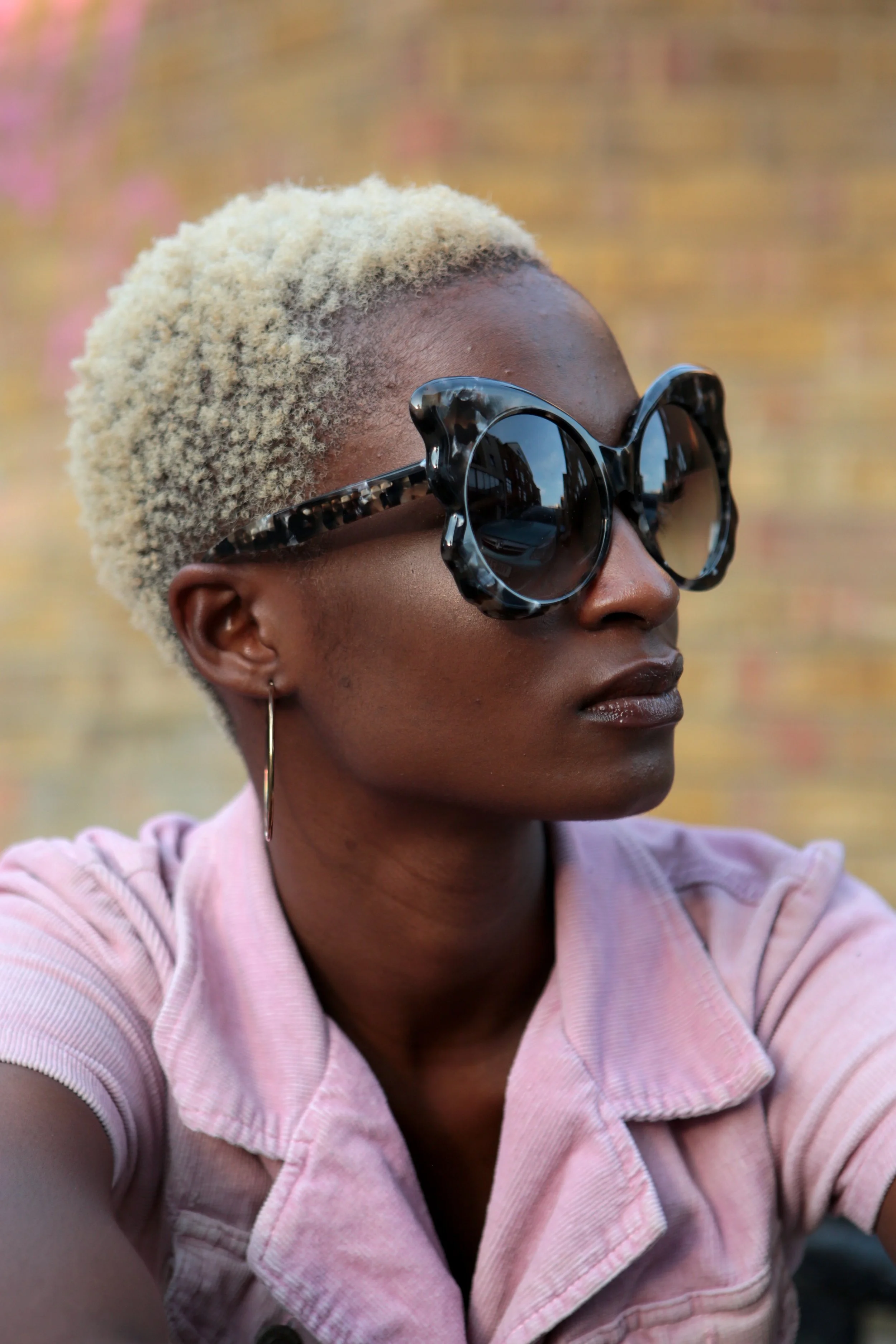 A woman with short, blonde curly hair wearing large, dark sunglasses, gold hoop earrings, and a light pink top, looking to the side.