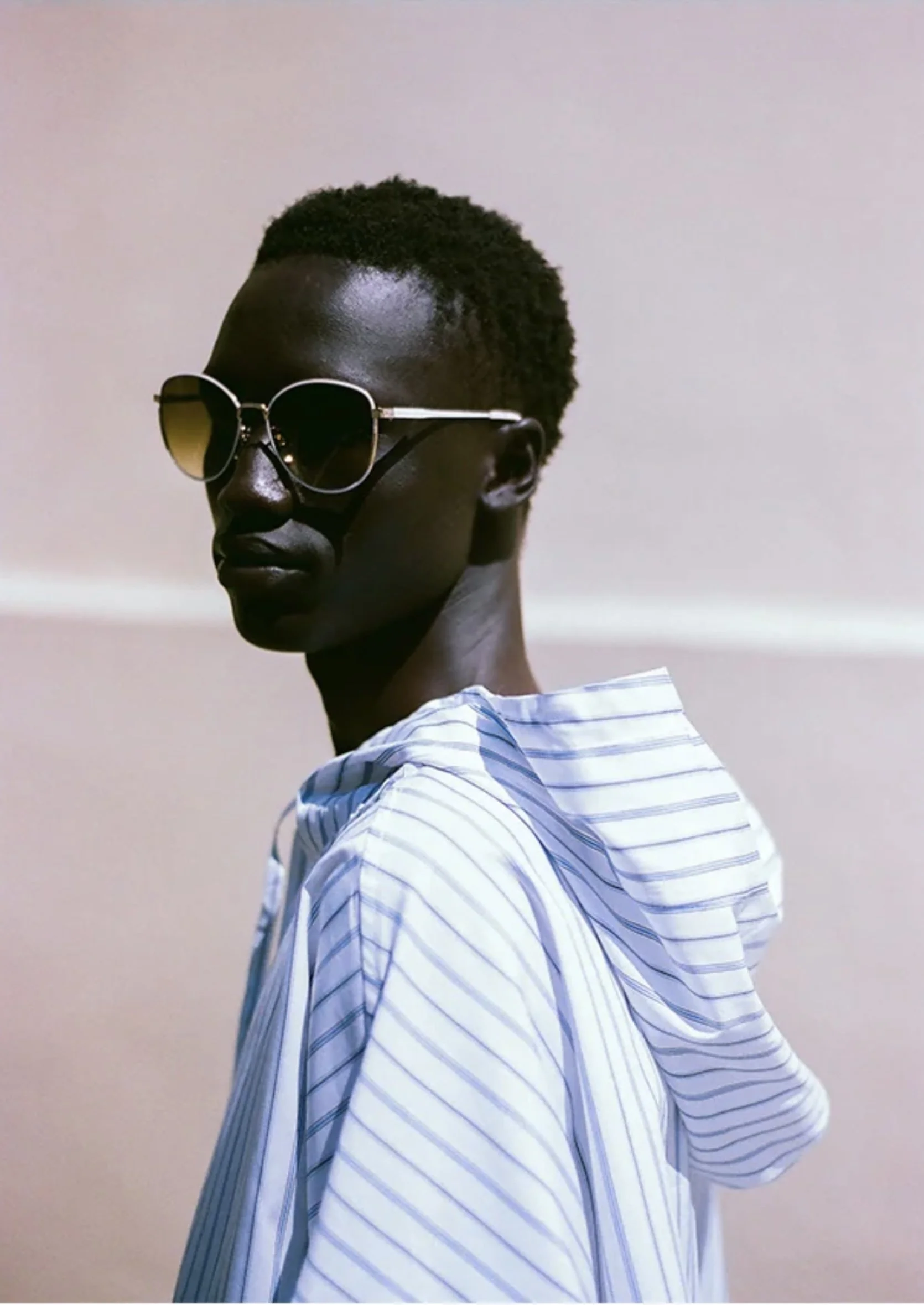 A young man with dark skin, wearing sunglasses and a white striped hoodie, looks over his shoulder against a plain beige background.