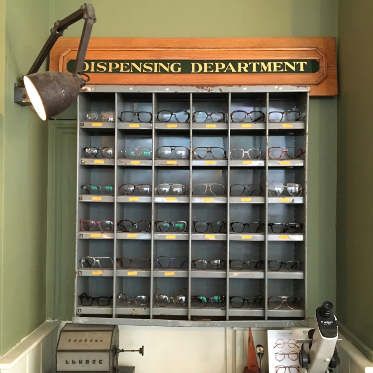 Metal display cabinet  full of eyewear in Specstacular Opticians, with a sign above that reads 'DISPENSING DEPARTMENT'.