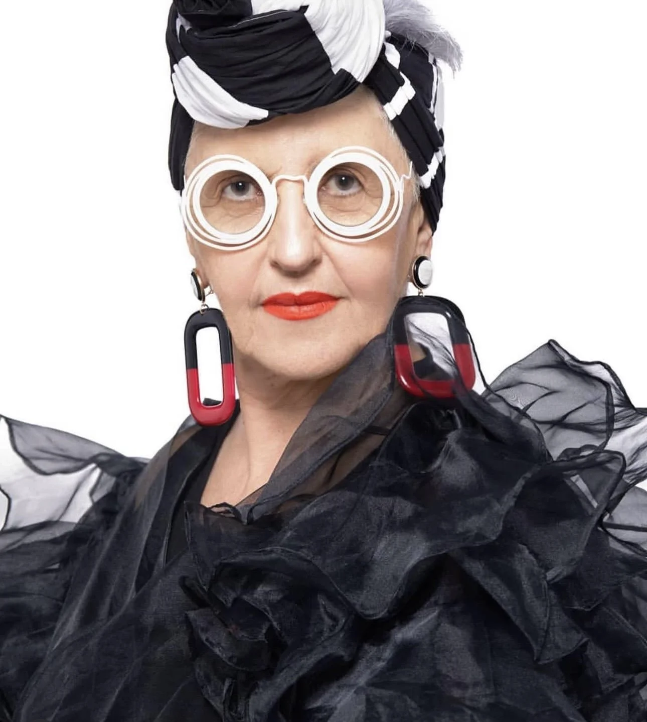 A woman wearing a black and white headscarf, round glasses, bright red lipstick, and large earrings with red and black accents. She has a sheer black garment with ruffled details.