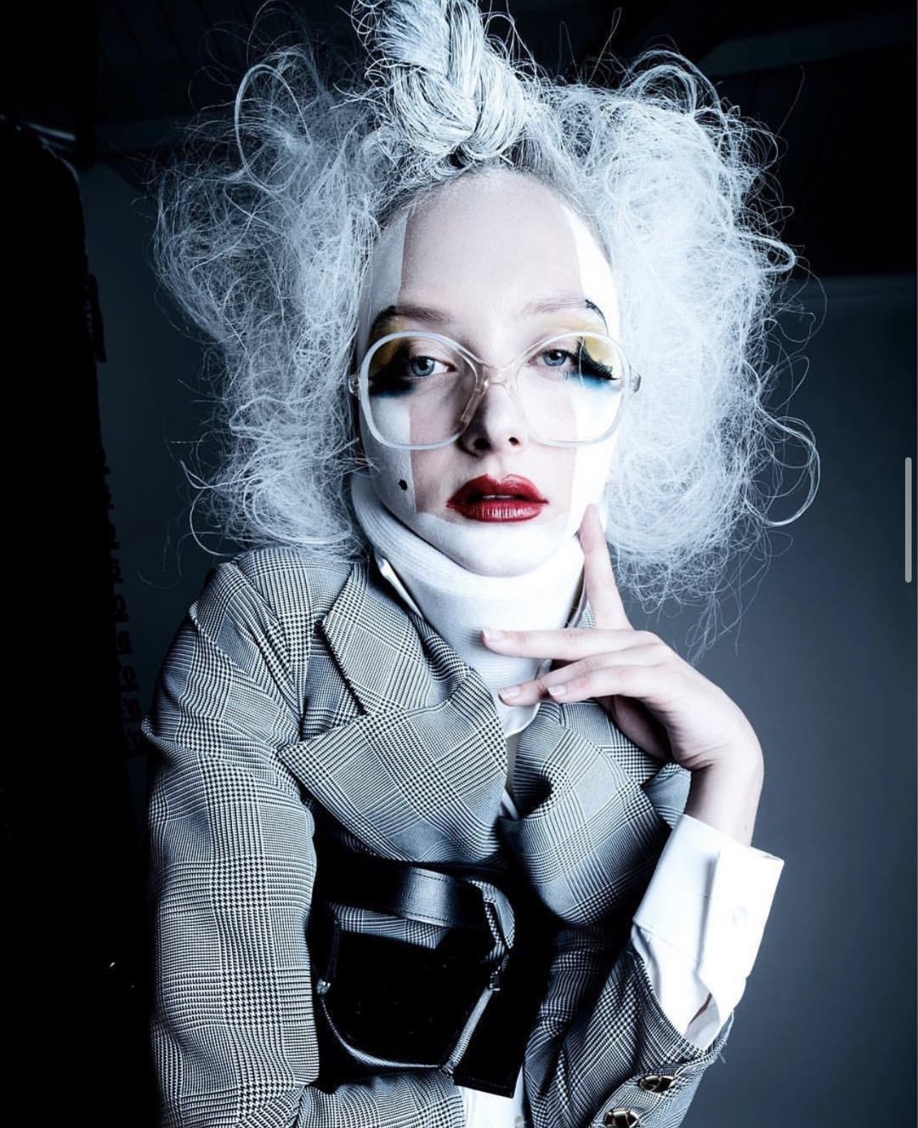 A woman with wild, curly gray hair, wearing large glasses, bright red lipstick, a plaid blazer, and a white turtleneck, posing with her hand near her face