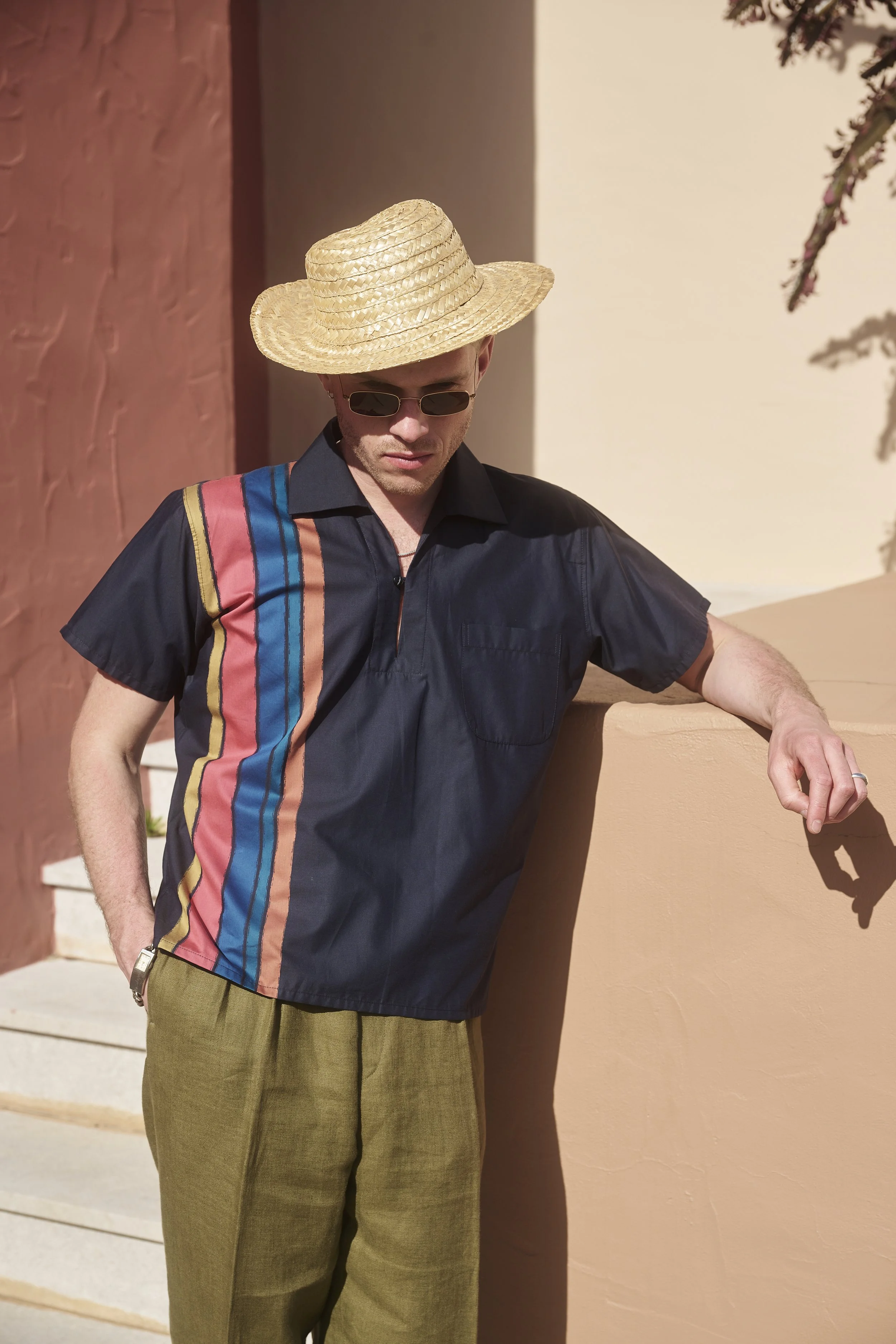 A man wearing sunglasses, a straw hat, a dark short-sleeve shirt with colorful stripes, and olive green pants, leaning against a peach-colored wall outdoors.