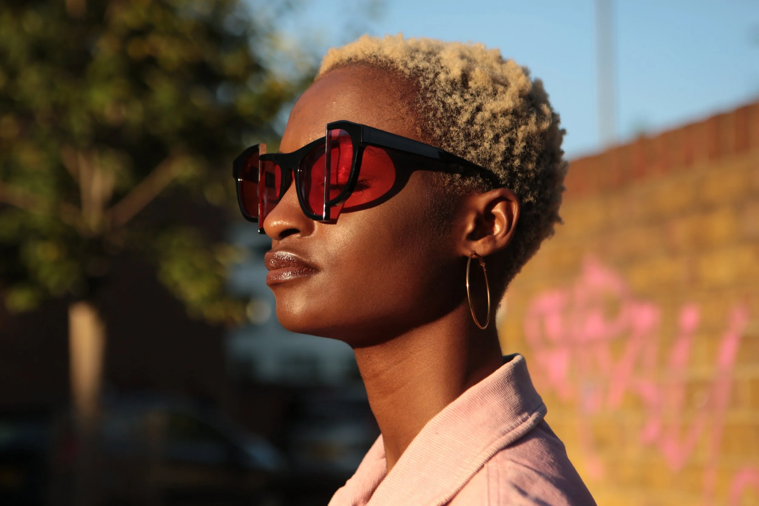 Close-up of a woman with short, blonde curly hair wearing large black sunglasses with red tinted lenses and hoop earrings, outdoors with a blurry background of trees and a brick wall.