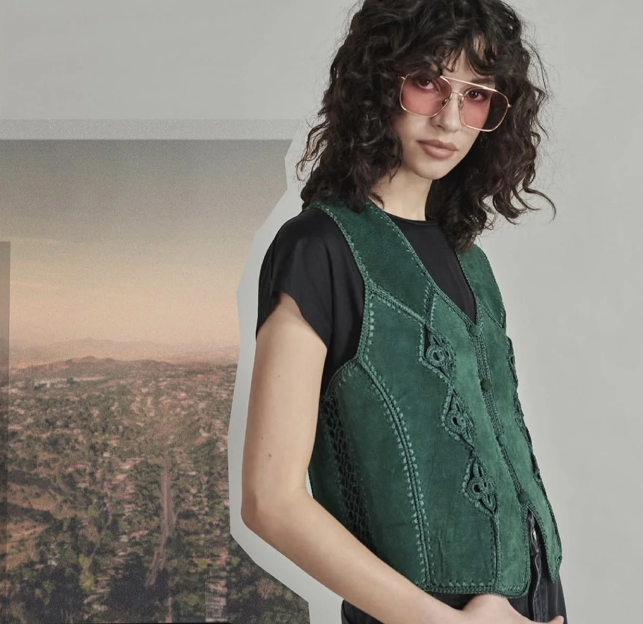 Young woman with curly dark hair wearing pink sunglasses and a black t-shirt, standing against a plain wall with a cityscape background image.