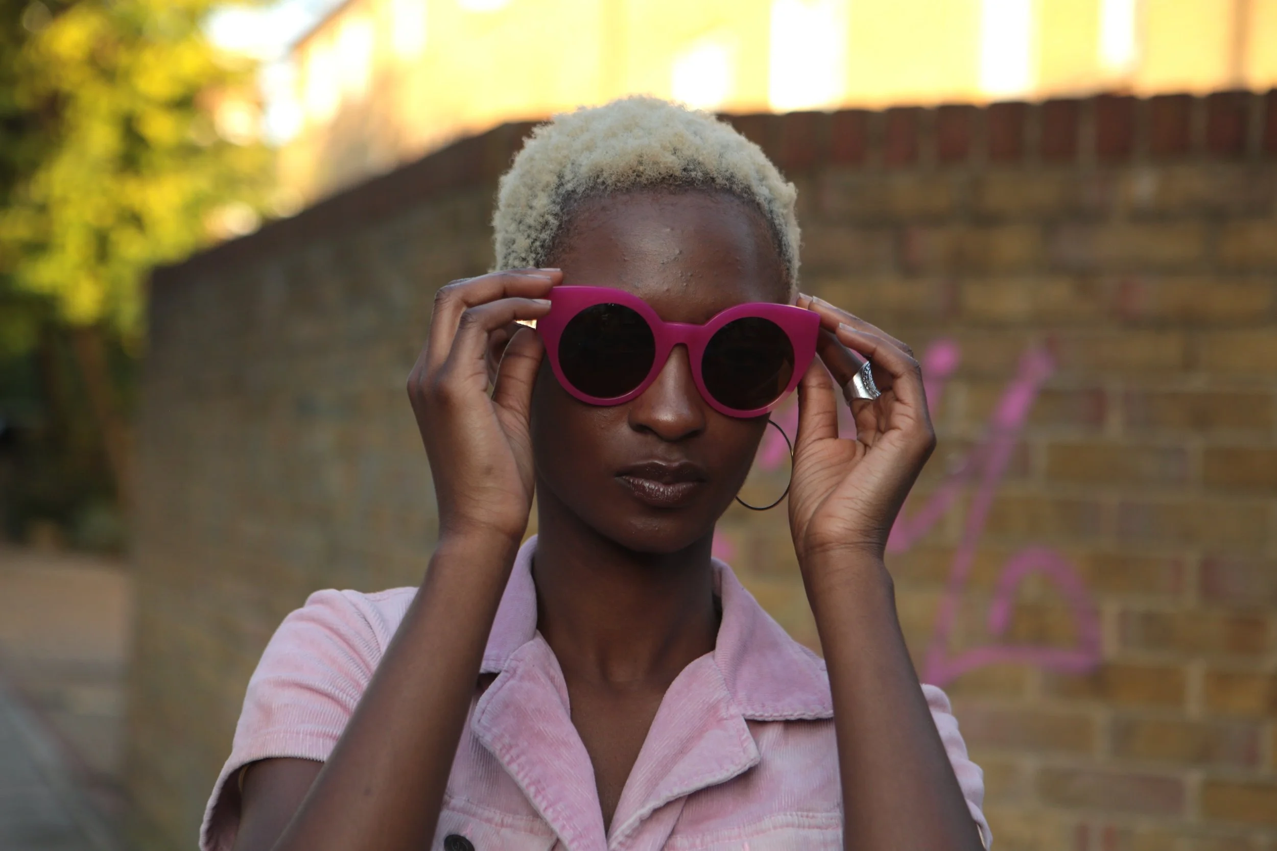 A woman with blonde hair wearing pink sunglasses adjusting them while standing outdoors in front of a brick wall.