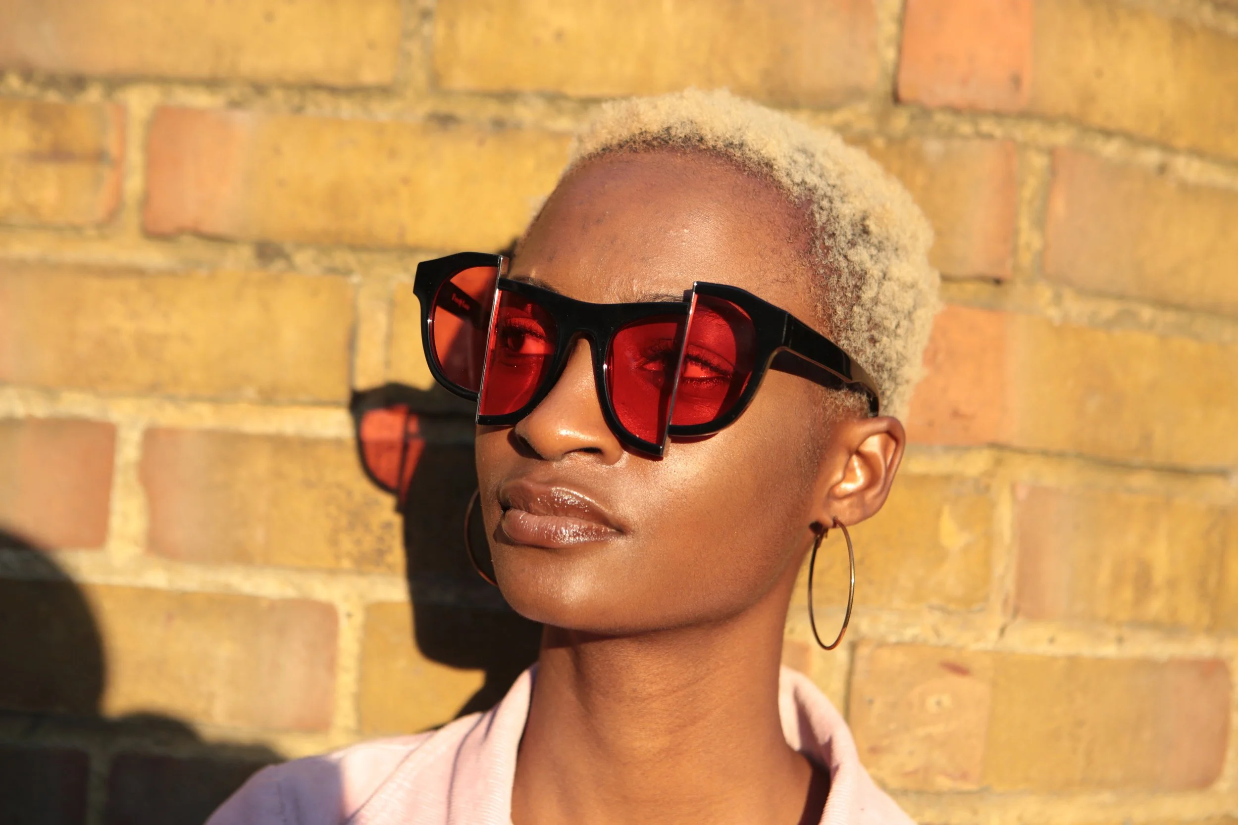 A woman with short, bleached blonde hair wearing large black sunglasses with red lenses, hoop earrings, and a light pink shirt, standing against a brick wall with sunlight casting a shadow.