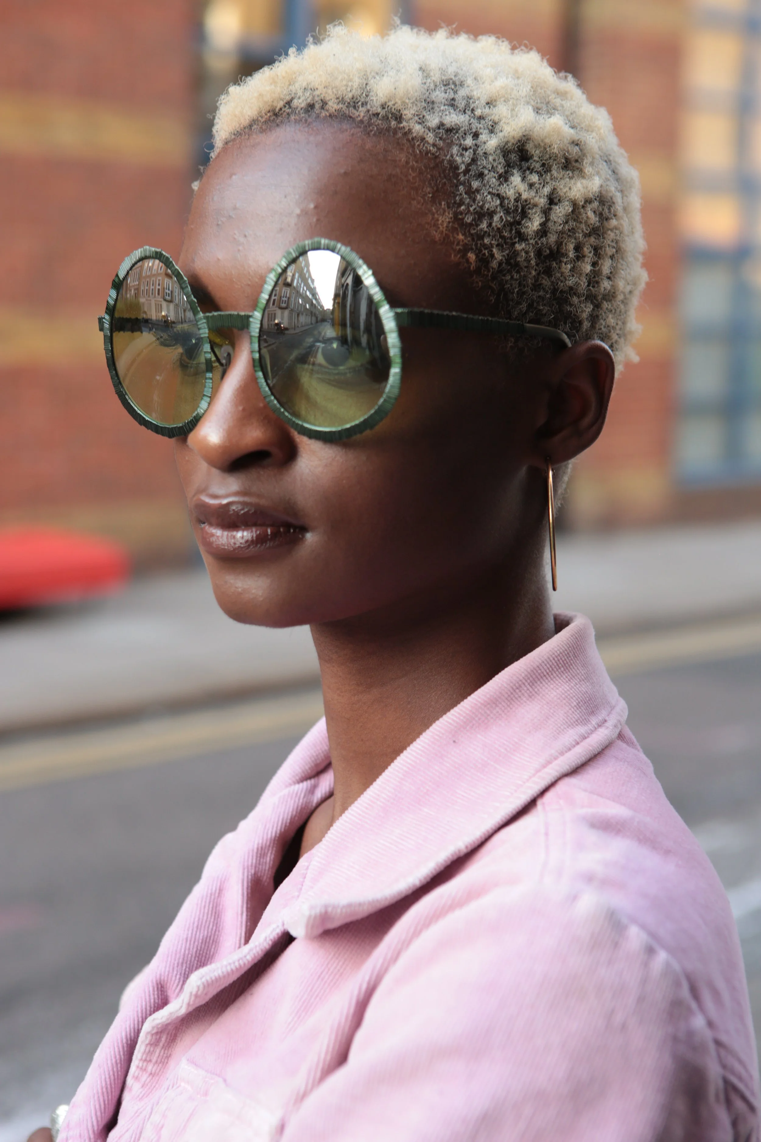A woman with short, bleached blonde hair wearing large, round sunglasses with a green textured frame, a pink jacket, and a gold hoop earring, standing outside in an urban setting with brick buildings in the background.