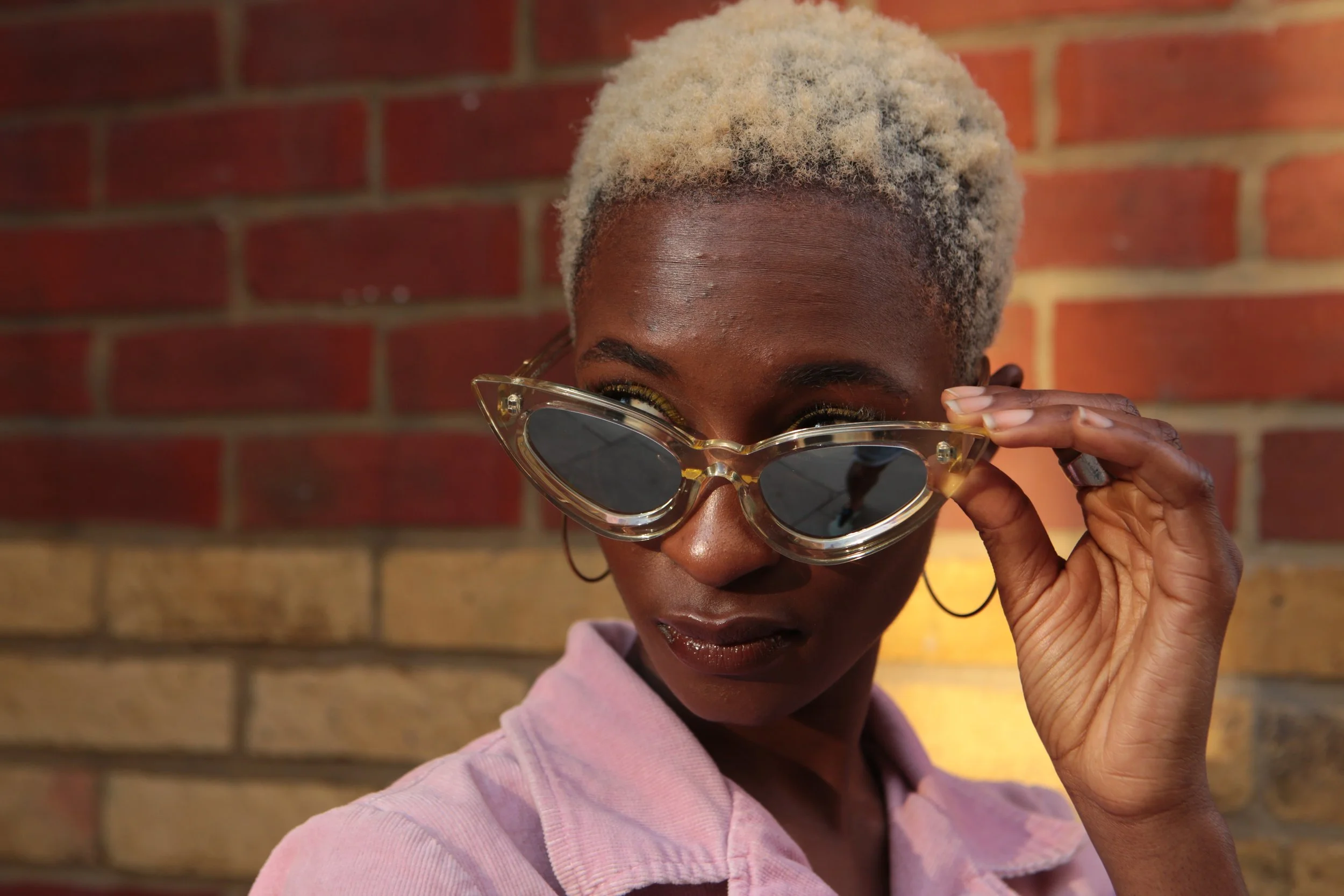A woman with short platinum blonde hair wearing a pink shirt, holding stylish transparent sunglasses with dark lenses near her eyes, standing against a brick wall background.
