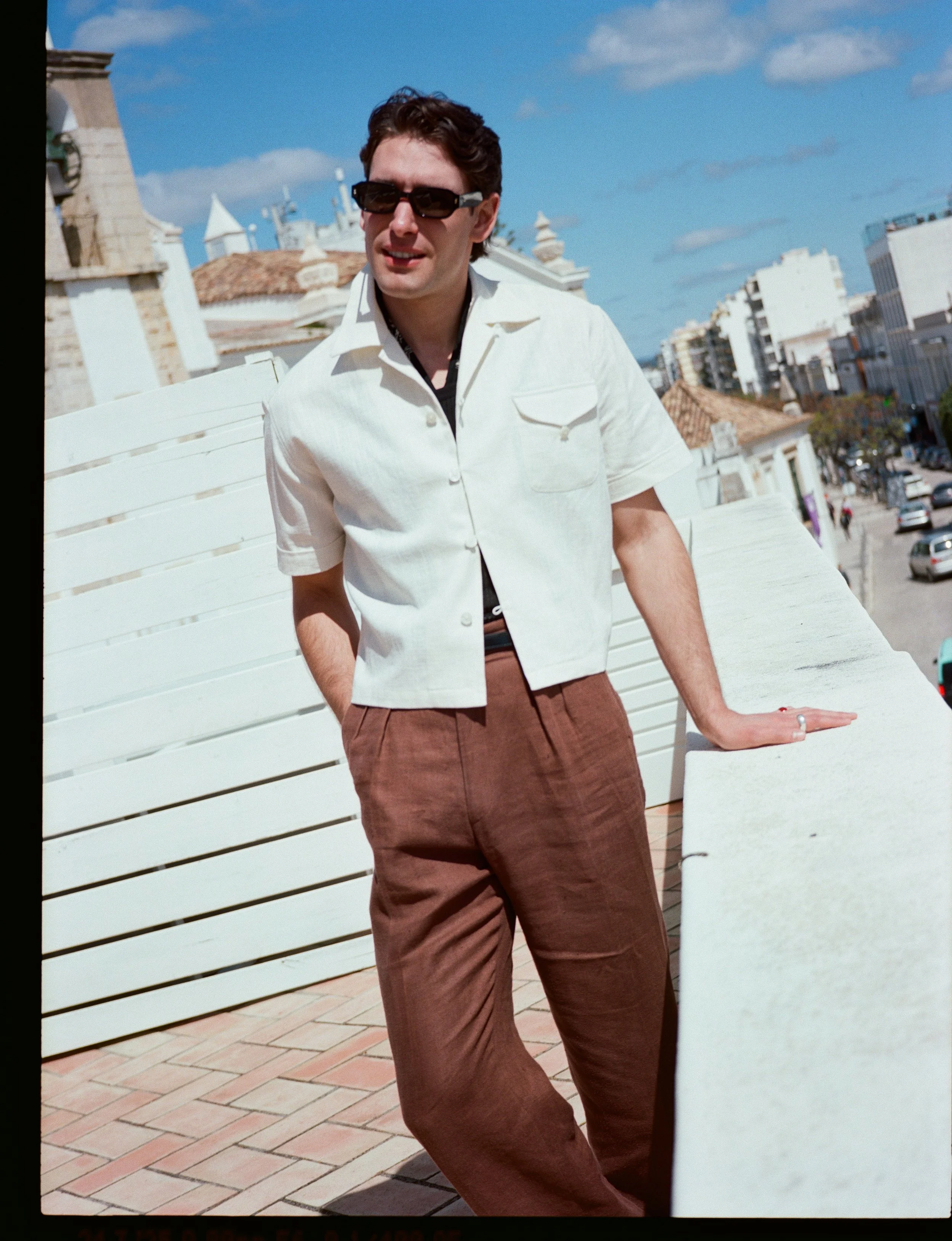 A young man stands outdoors on a sunny day wearing sunglasses, a white short-sleeve shirt, and brown pants, leaning with one hand on a white ledge with a cityscape in the background.