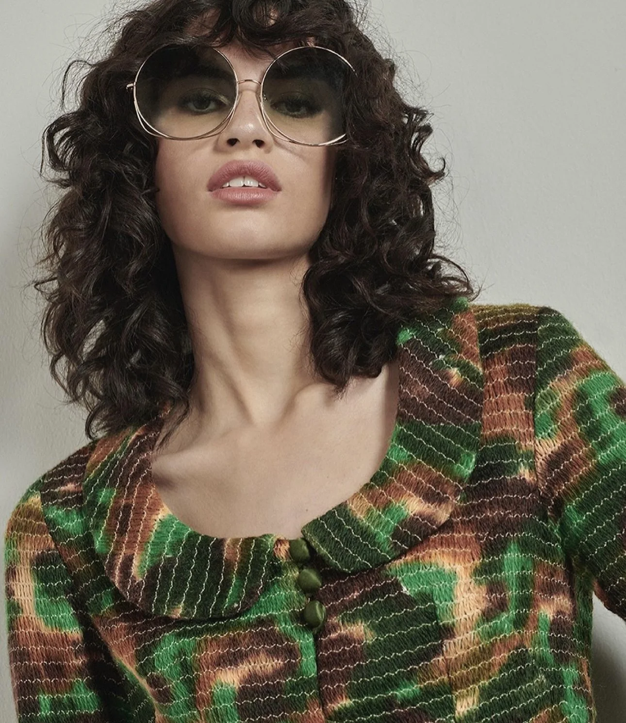 A woman with curly dark hair, wearing large round sunglasses, and a colorful patterned jacket with a rounded collar and green buttons.