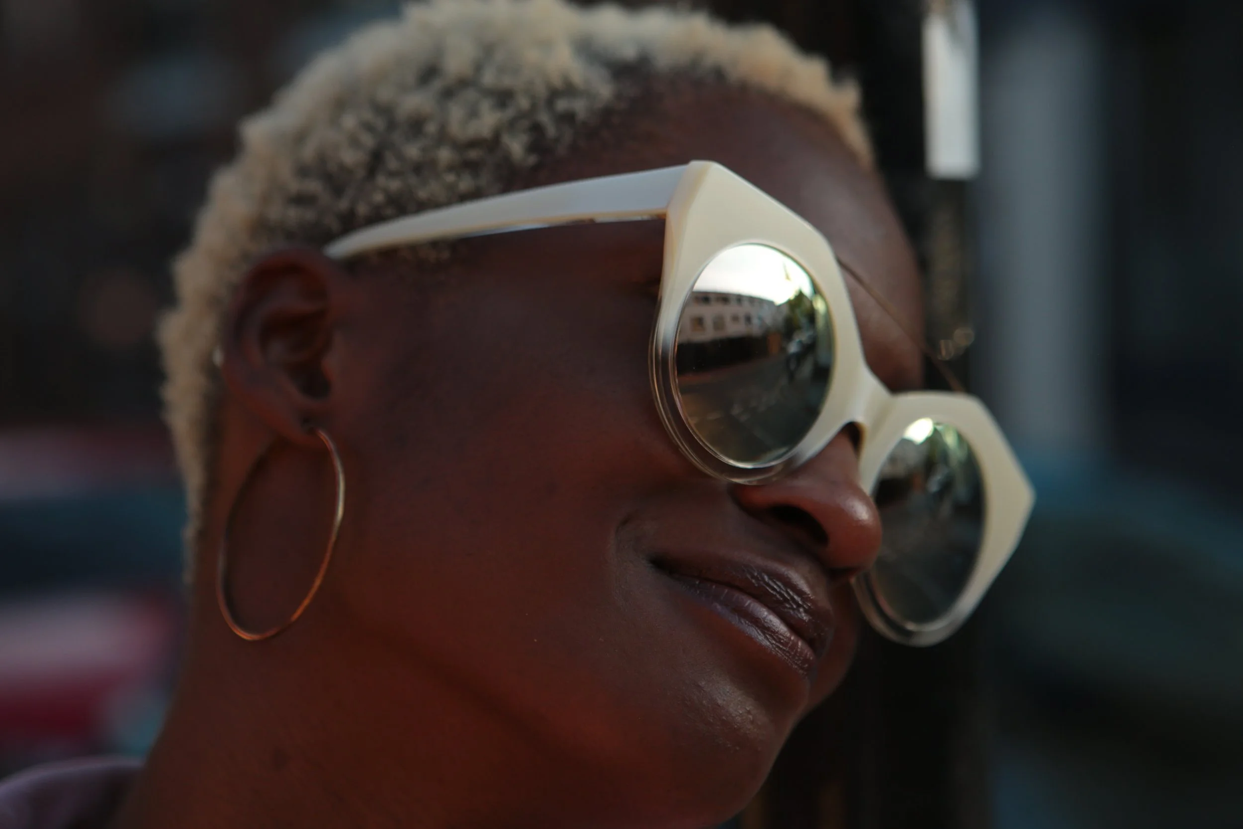 Close-up of a woman with short blonde hair, wearing large cream-colored sunglasses and hoop earrings, smiling with her eyes closed.
