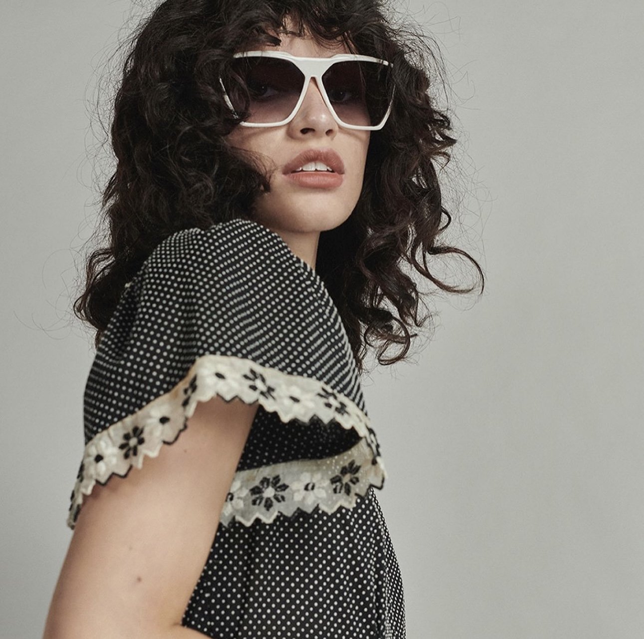 A woman with curly dark hair wearing large white sunglasses and a black dress with white polka dots and floral lace trim on the sleeve, looking slightly to the side against a plain background.