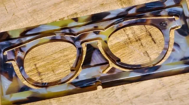 Close-up of a sheet tortoiseshell pattern acetate with hand cut shape of eyeglasses, placed on a wooden surface.