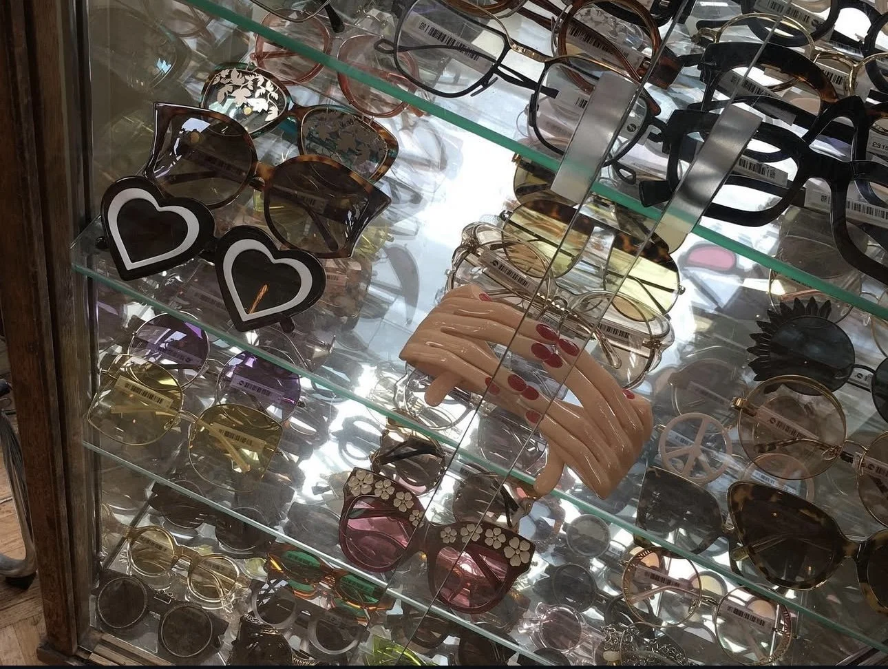 A glass display case filled with various sunglasses, including heart-shaped sunglasses, round, oval, and butterfly styles, with some decorative frames and a plastic hand model holding sunglasses.