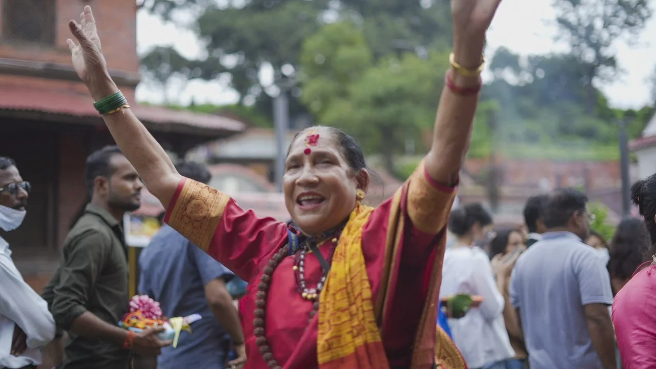 A woman in traditional colorful Indian attire with jewelry and a bindi on her forehead, smiling and raising her arms in celebration among a crowd outdoors.
