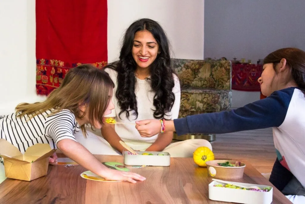 Priyanka Lugani smiling warmly while sharing a healthy meal with two young children. She is wearing a white sweater, sitting on the floor in a bright, conscious living setting. Priyanka is an advocate for children’s education and the founder of The G