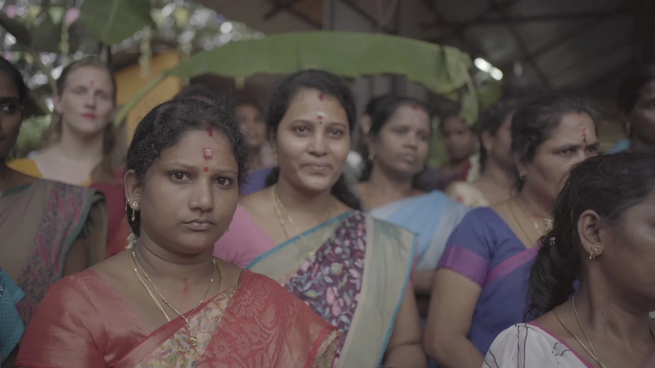 A group of women dressed in colorful traditional Indian sarees, standing outdoors under a roof with trees and sunlight in the background.