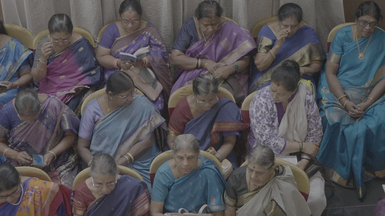 Group of Indian women dressed in traditional sarees seated closely together at an indoor event, some using mobile phones and reading.