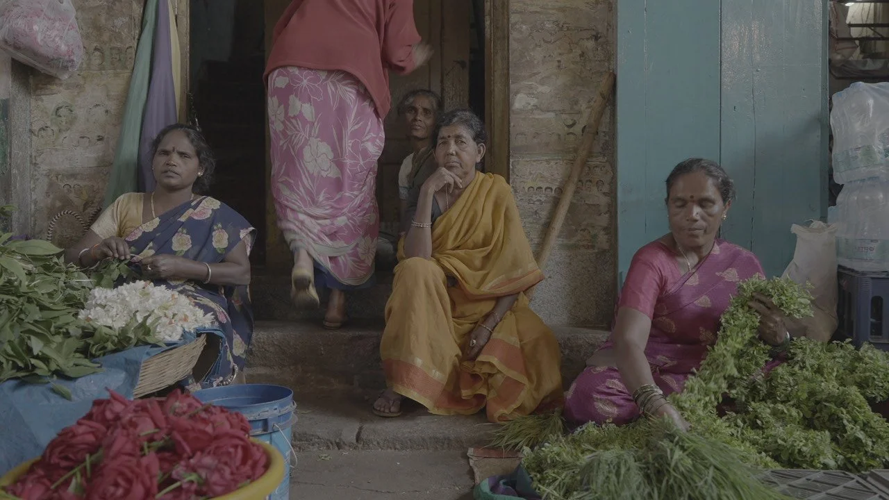 Women sitting outside a local market surrounded by flowers and greenery, some women are holding flowers while seated.