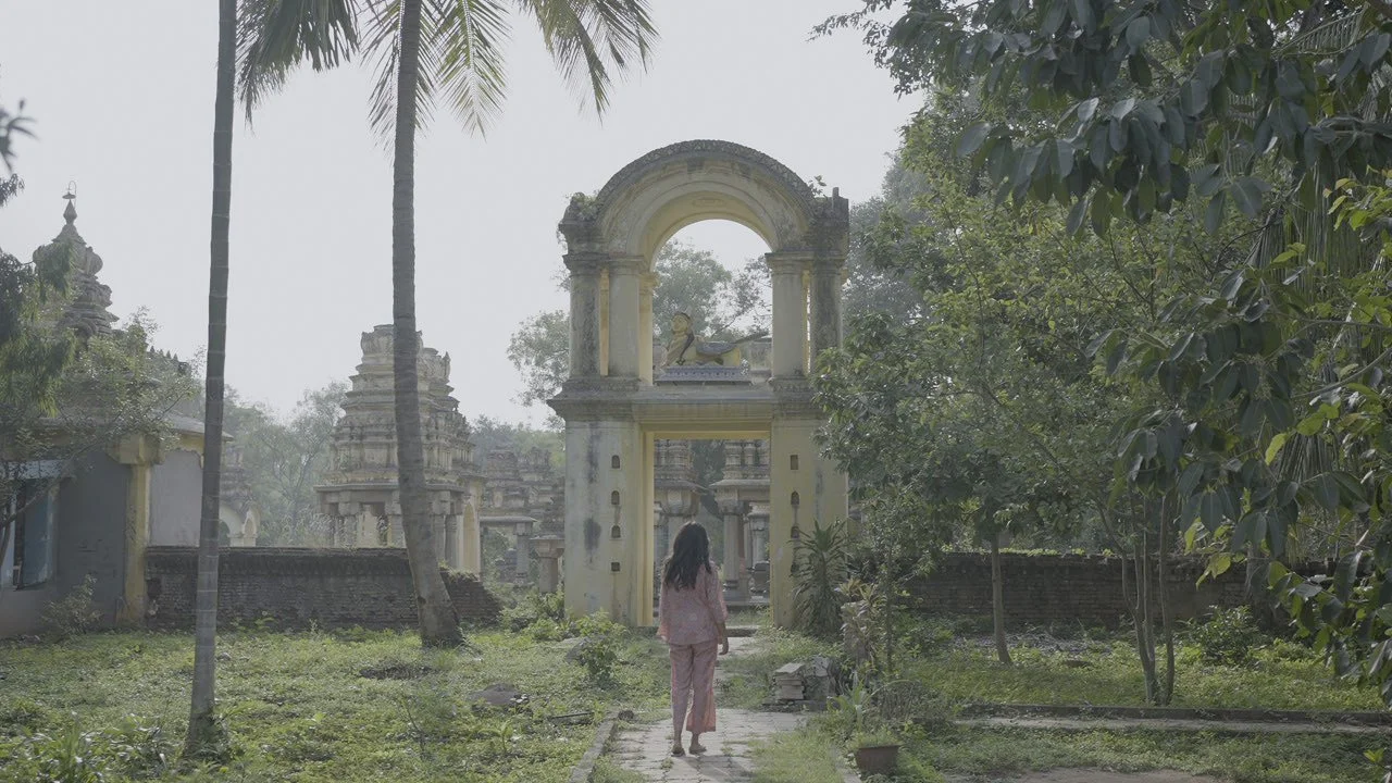 A girl in pink clothes walking down a stone pathway toward an ornate archway, with old temple structures and trees on either side, in a peaceful outdoor setting.