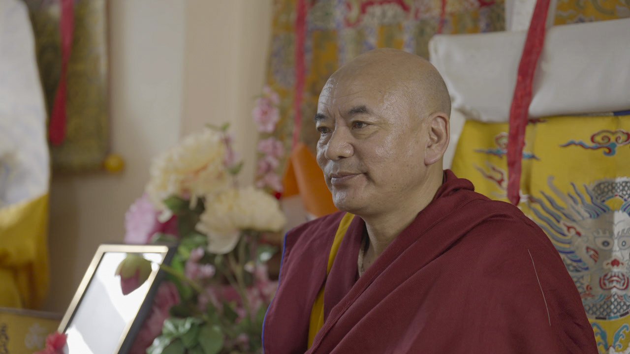 A Buddhist monk in maroon robes sitting inside a temple surrounded by colorful textiles, flowers, and traditional decorations.