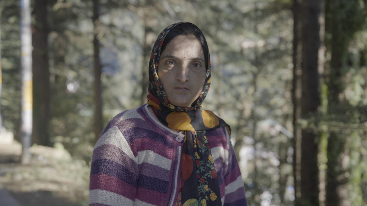 A woman with a colorful headscarf and striped sweater standing outdoors in a forested area.