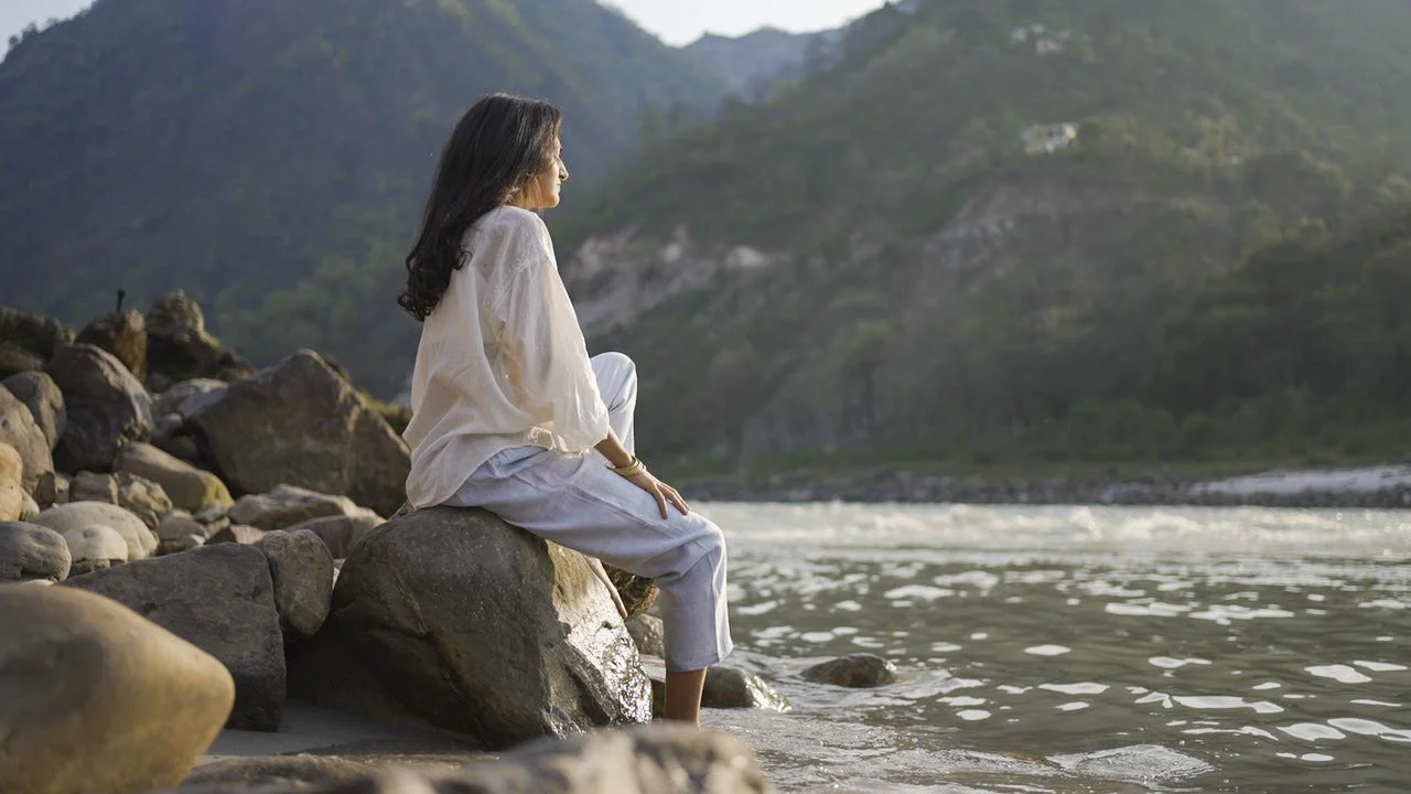 Priyanka Lugani sitting on riverside rocks by the Ganga in Rishikesh, looking toward the mountains-
