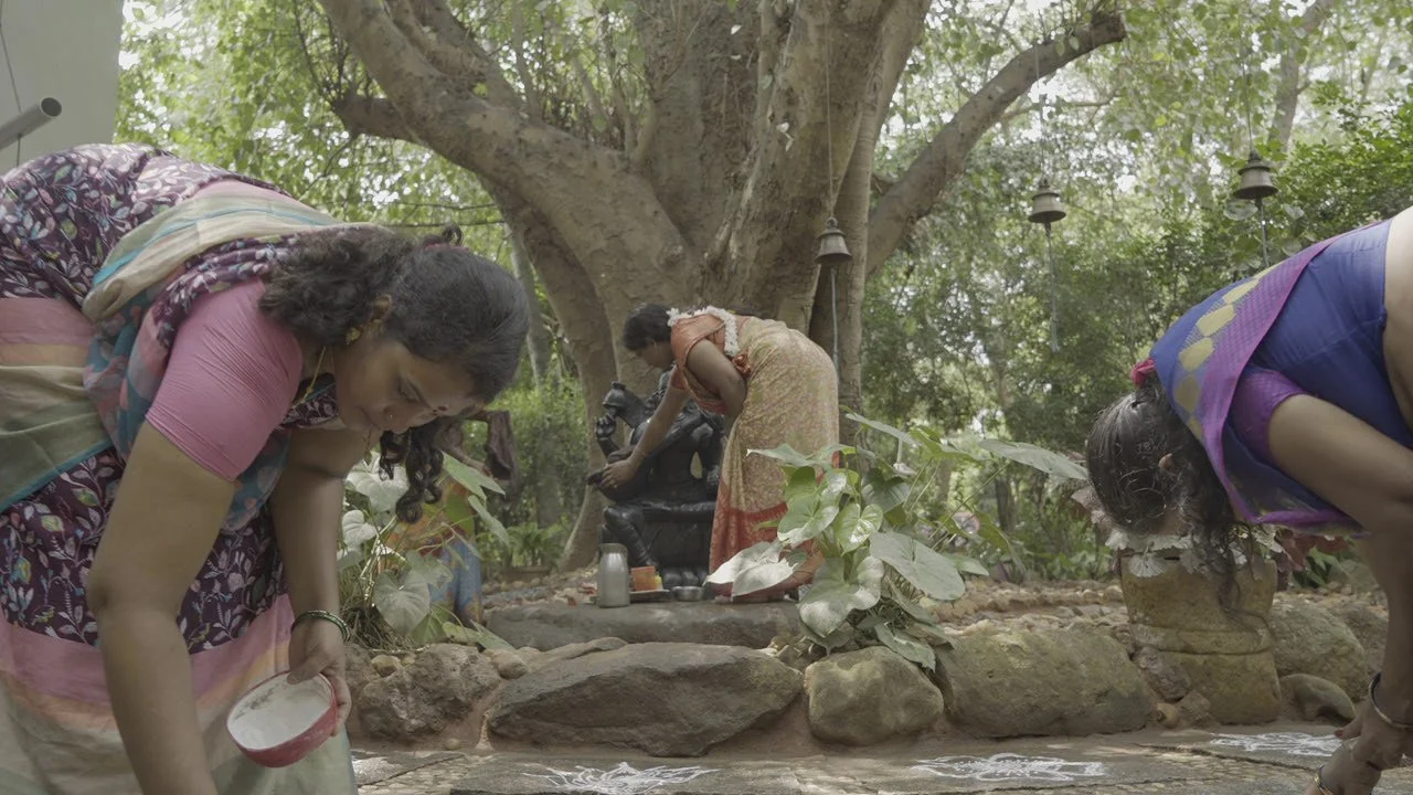 Three women dressed in traditional Indian sarees are performing a ritual or cleansing ceremony by a water body outdoors, with a large tree and hanging bells in the background.