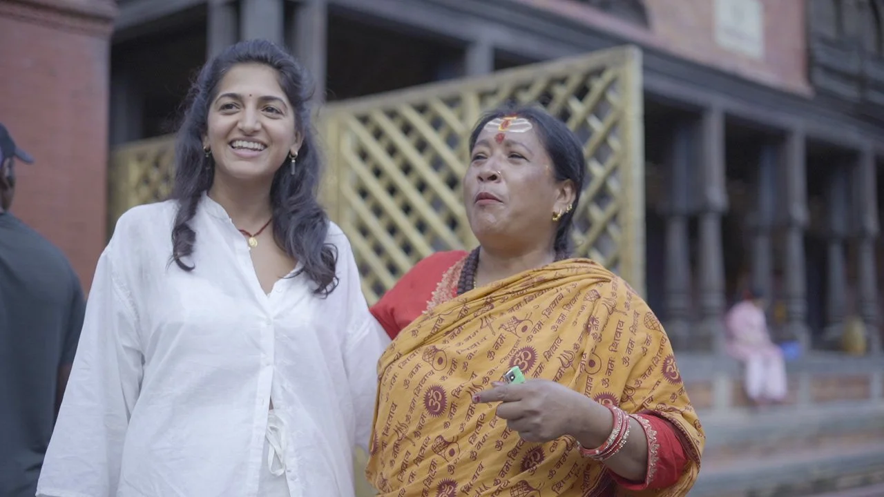 Two women, one wearing a white shirt and the other wearing a yellow and red sari, smiling and standing outside near a building with wooden support beams.