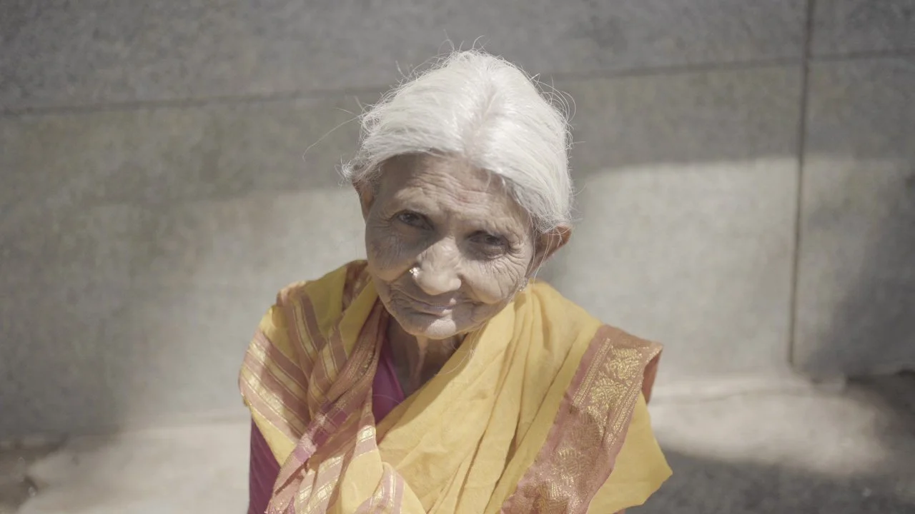 A close-up of an elderly woman with white hair, wearing a traditional yellow and pink saree, standing outdoors against a stone wall background.