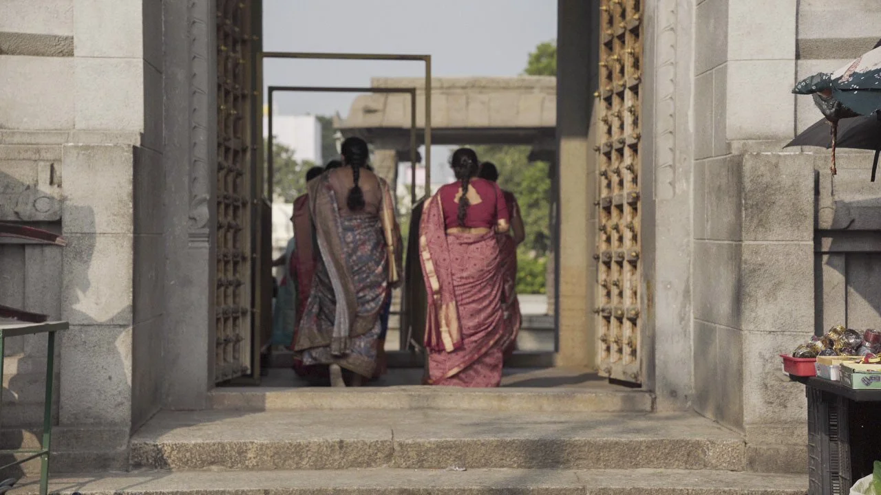 Women wearing traditional colorful sarees walking through a large stone temple gateway into a sunny courtyard.