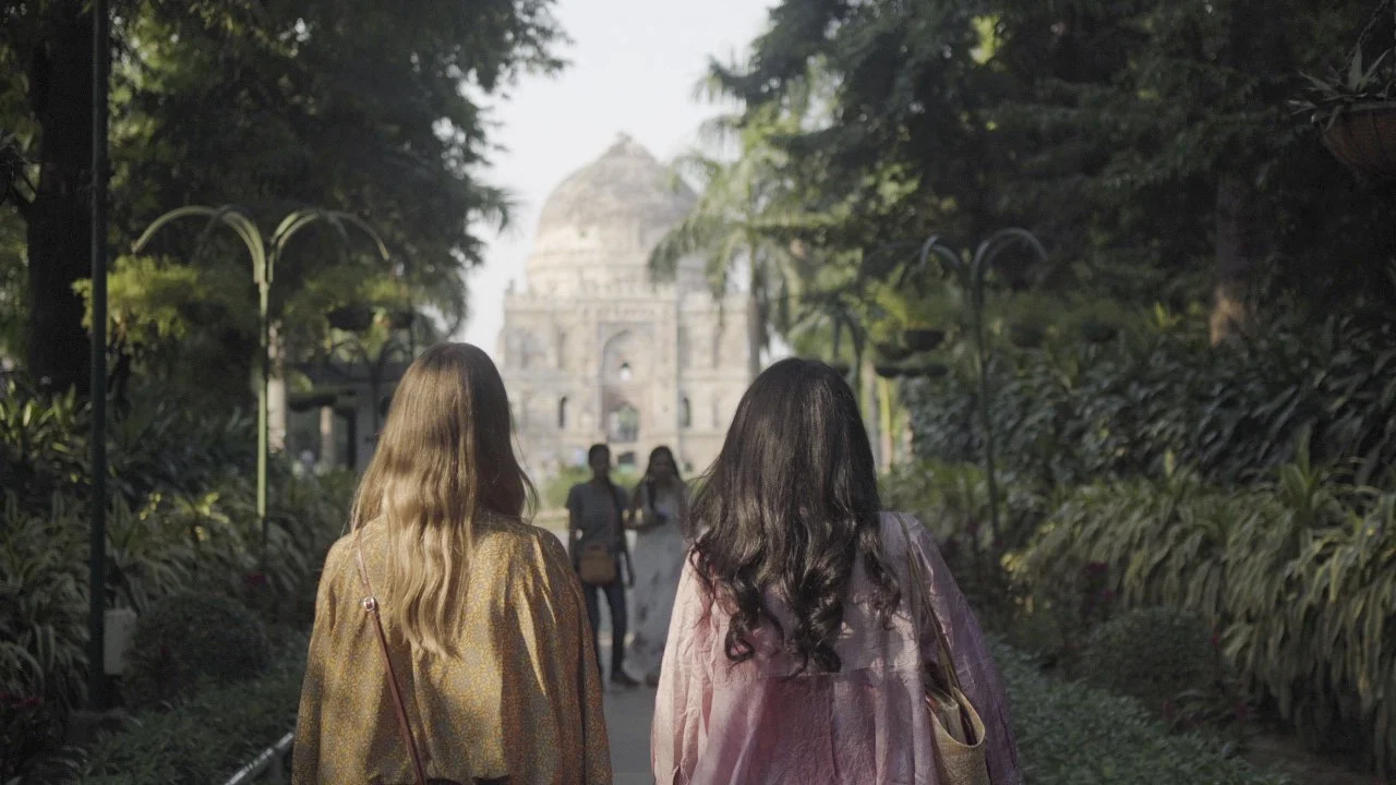Two women walking through a lush garden towards a historic monument, the Taj Mahal, in India.