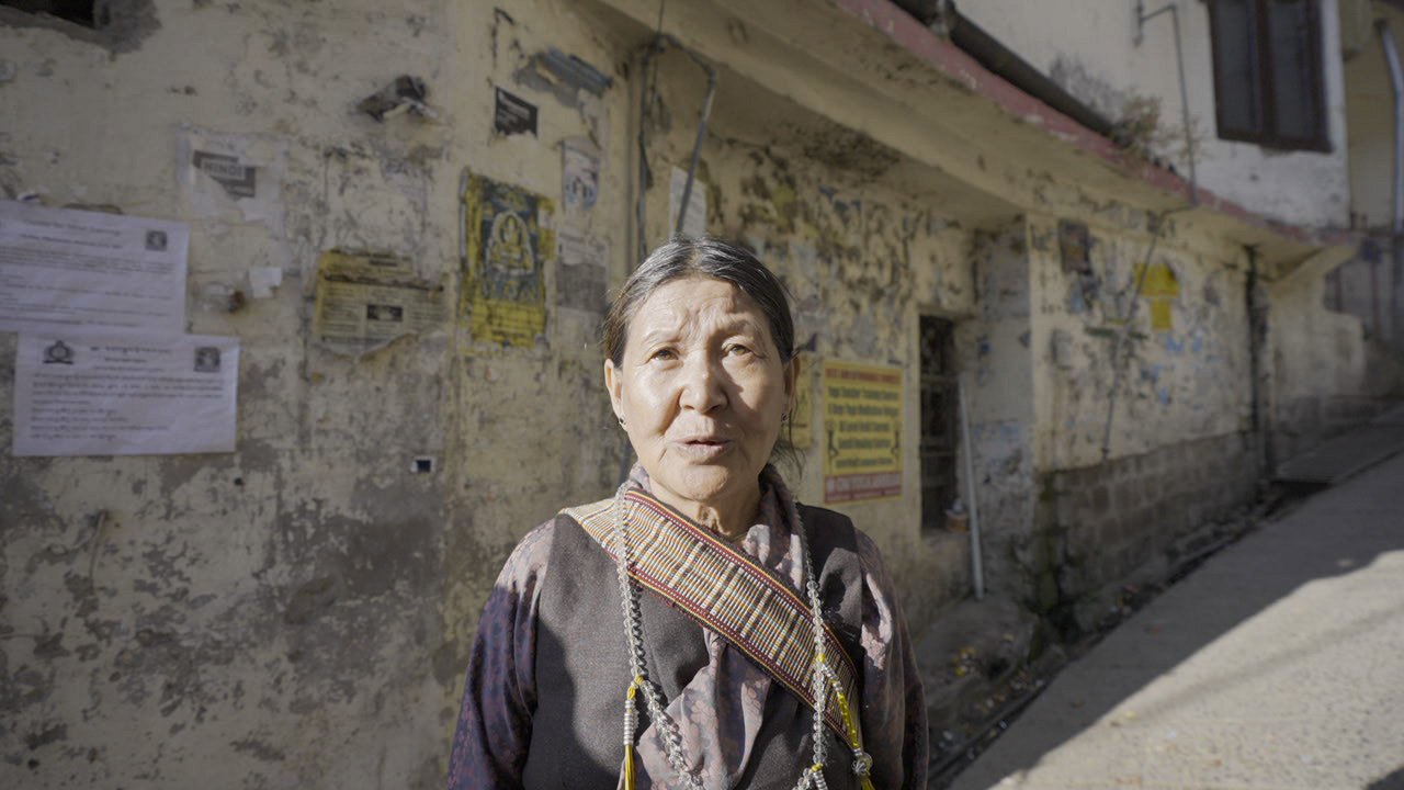 An elderly woman with traditional attire standing outside a weathered building with posters on the wall.