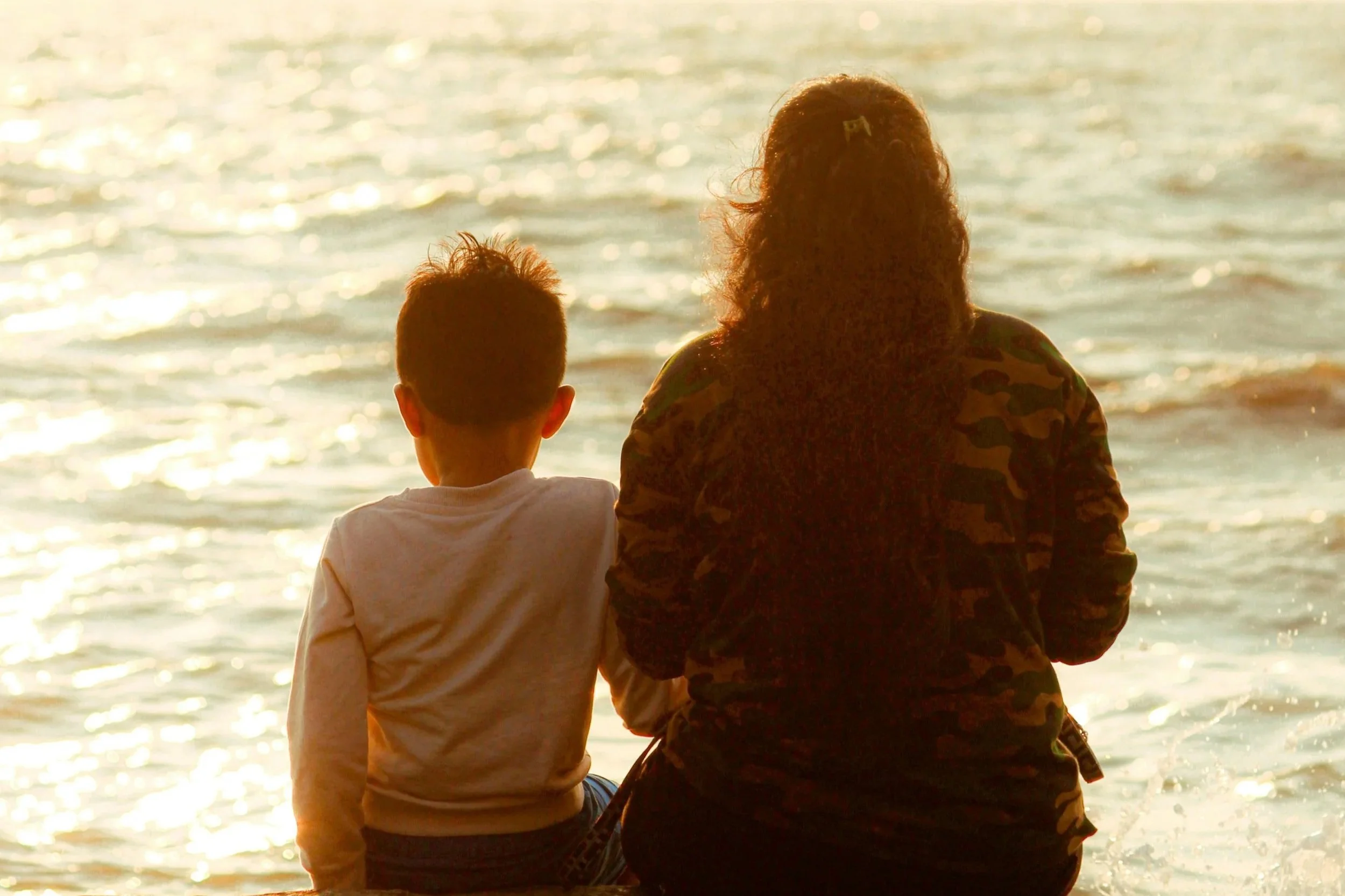 A woman and child sitting together by the ocean at sunset, watching the waves in a quiet moment of connection and reflection.