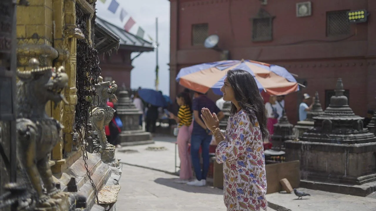 Priyanka Lugani in a prayer position (Anjali Mudra) at a temple in Nepal. She is wearing a floral patterned top and gold bangles, standing before an ornate golden shrine with stone lion statues and prayer flags in the background.