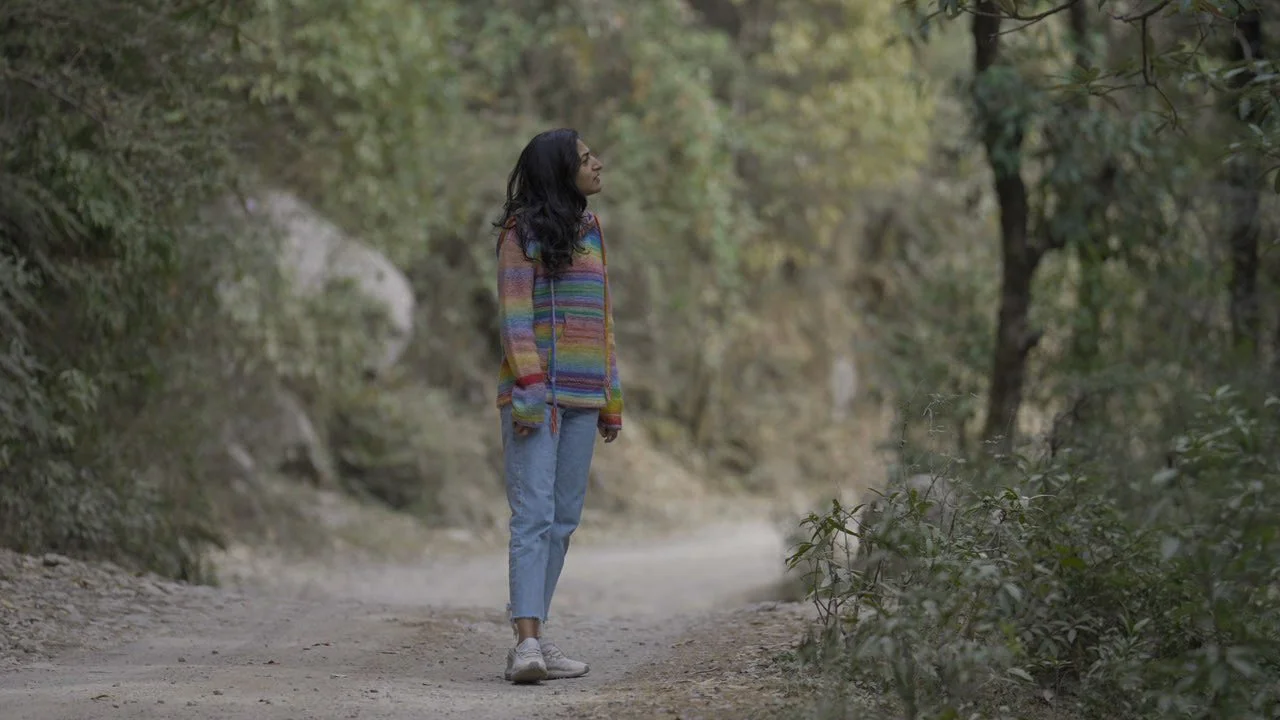 A young woman with long dark hair, wearing a multicolored sweater and light blue jeans, stands on a forest trail surrounded by trees and bushes.