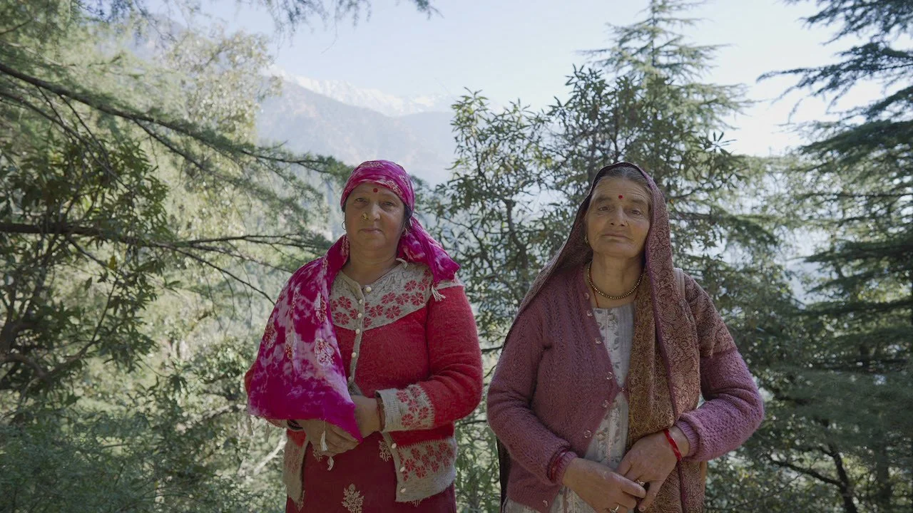 Two older women standing outdoors among trees with mountains in the background. They are dressed in traditional clothing and have serious expressions.