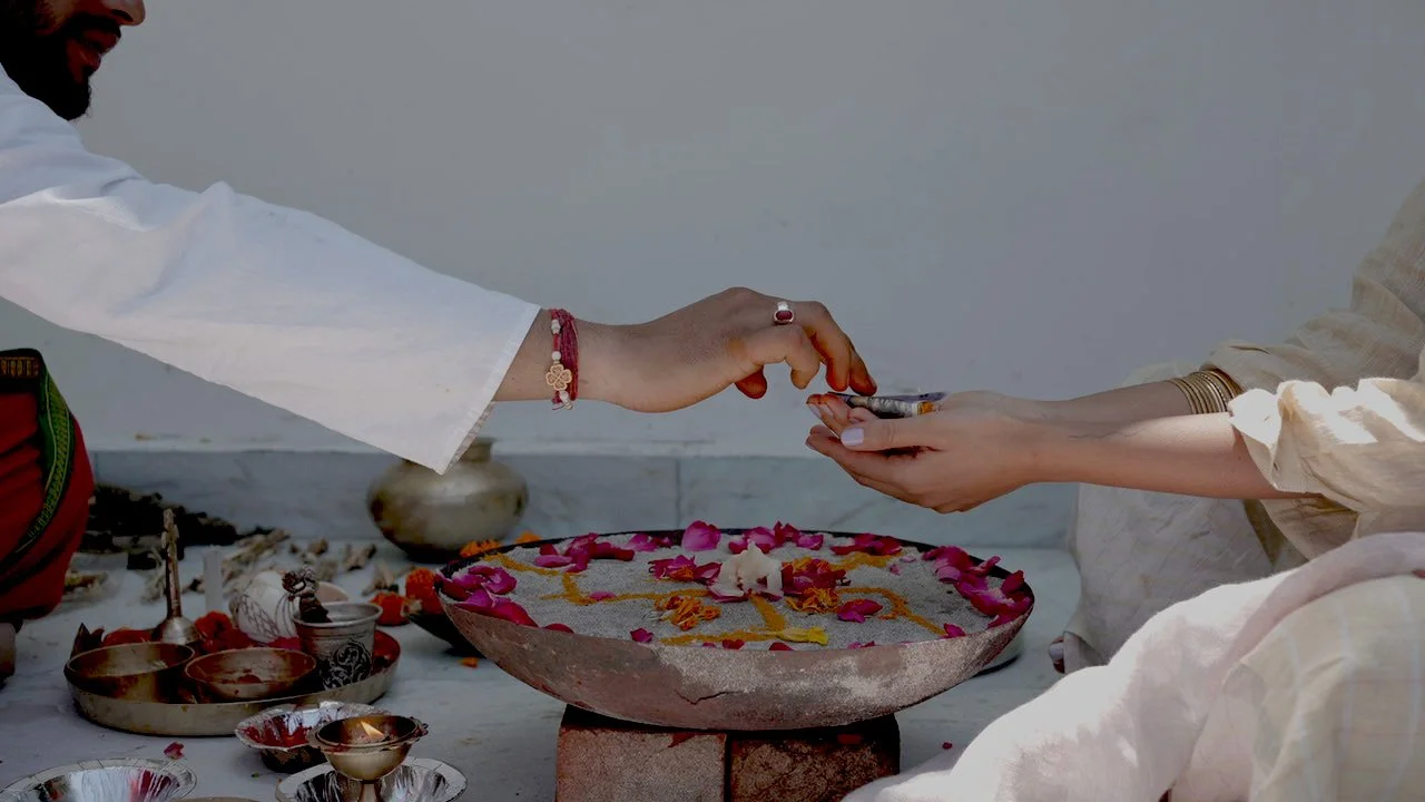 Person transferring a small object, possibly a gift or offering, to another person's hand during a religious or spiritual ceremony, with flower petal decorations and traditional items on a tray.