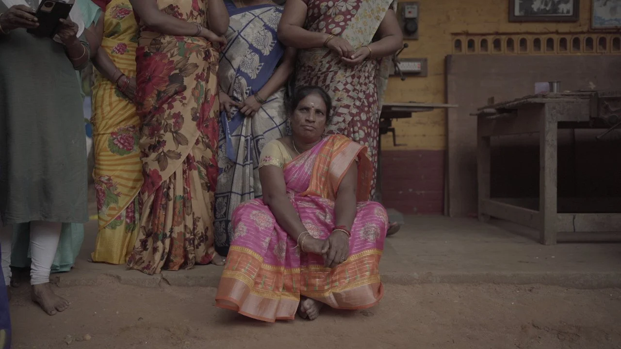A woman sitting on the ground among a group of women standing behind her, inside a rustic room with wooden walls and a work table.