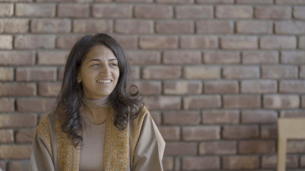Priyanka Lugani, founder of The Girl with the Goddess, smiling in a brown patterned shawl against a brick wall in Dharamshala.