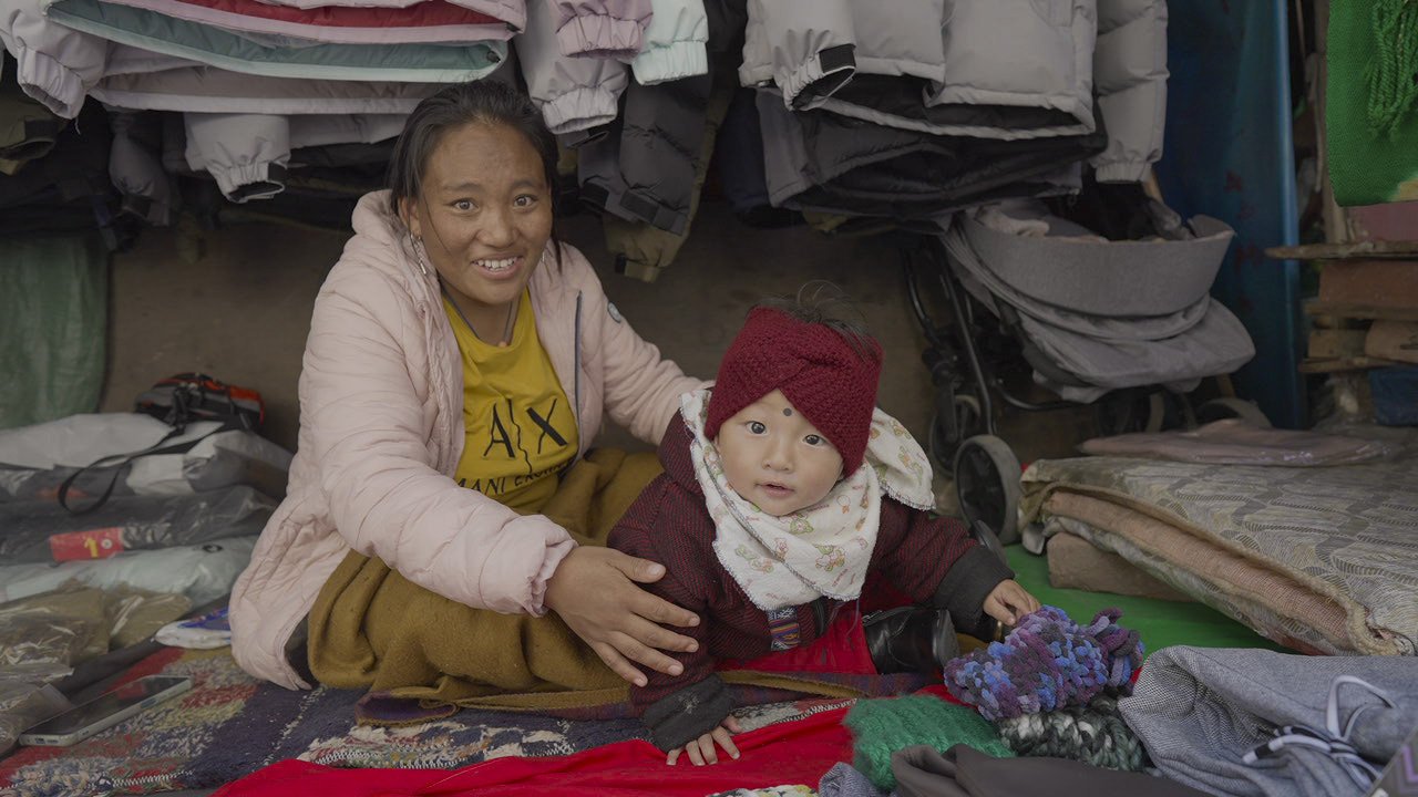 A woman and a child sitting on the floor among clothing and blankets in a small space, smiling at the camera.