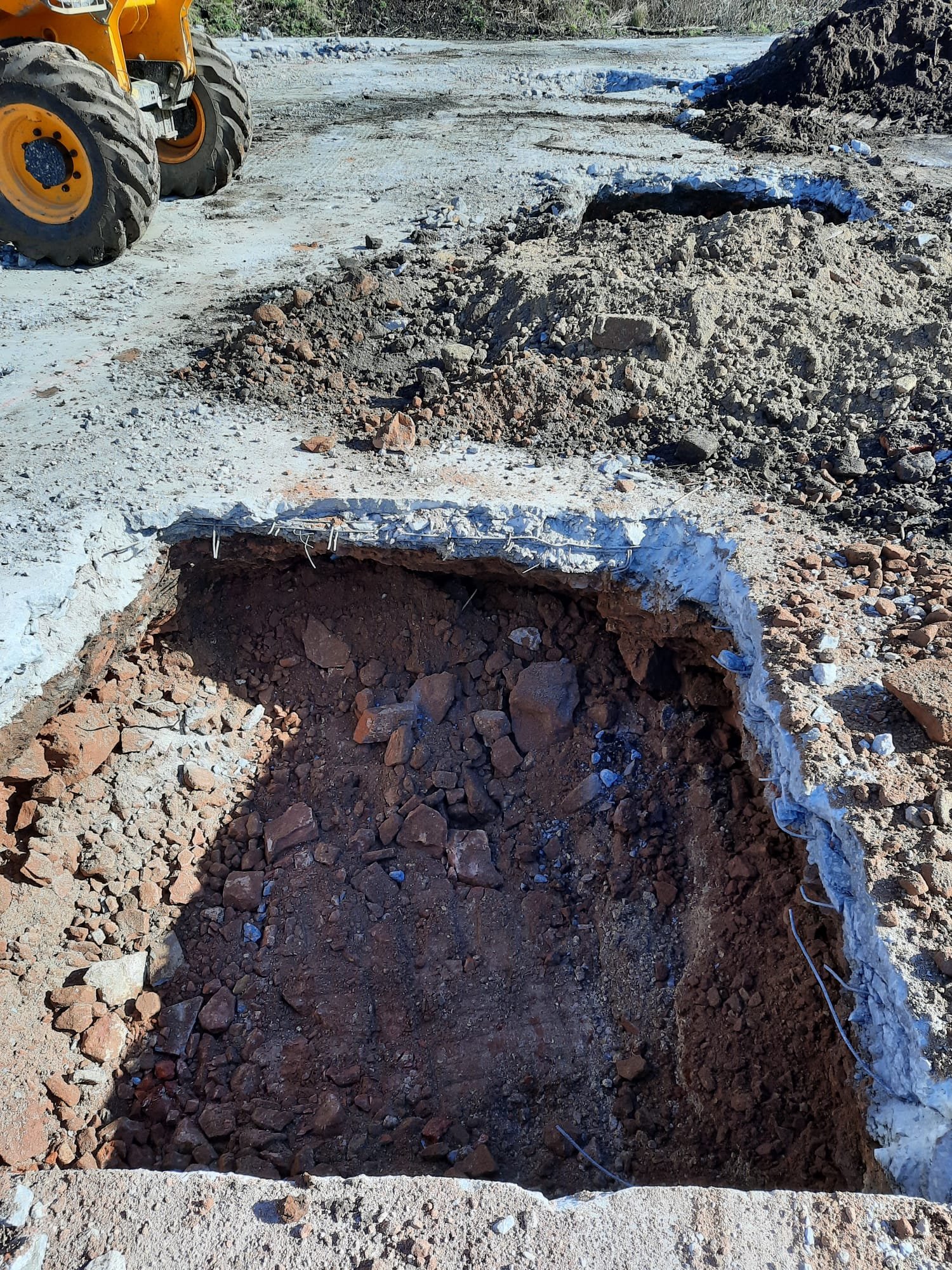 A construction site with a large hole in the ground and dirt piles, with a yellow backhoe in the background.