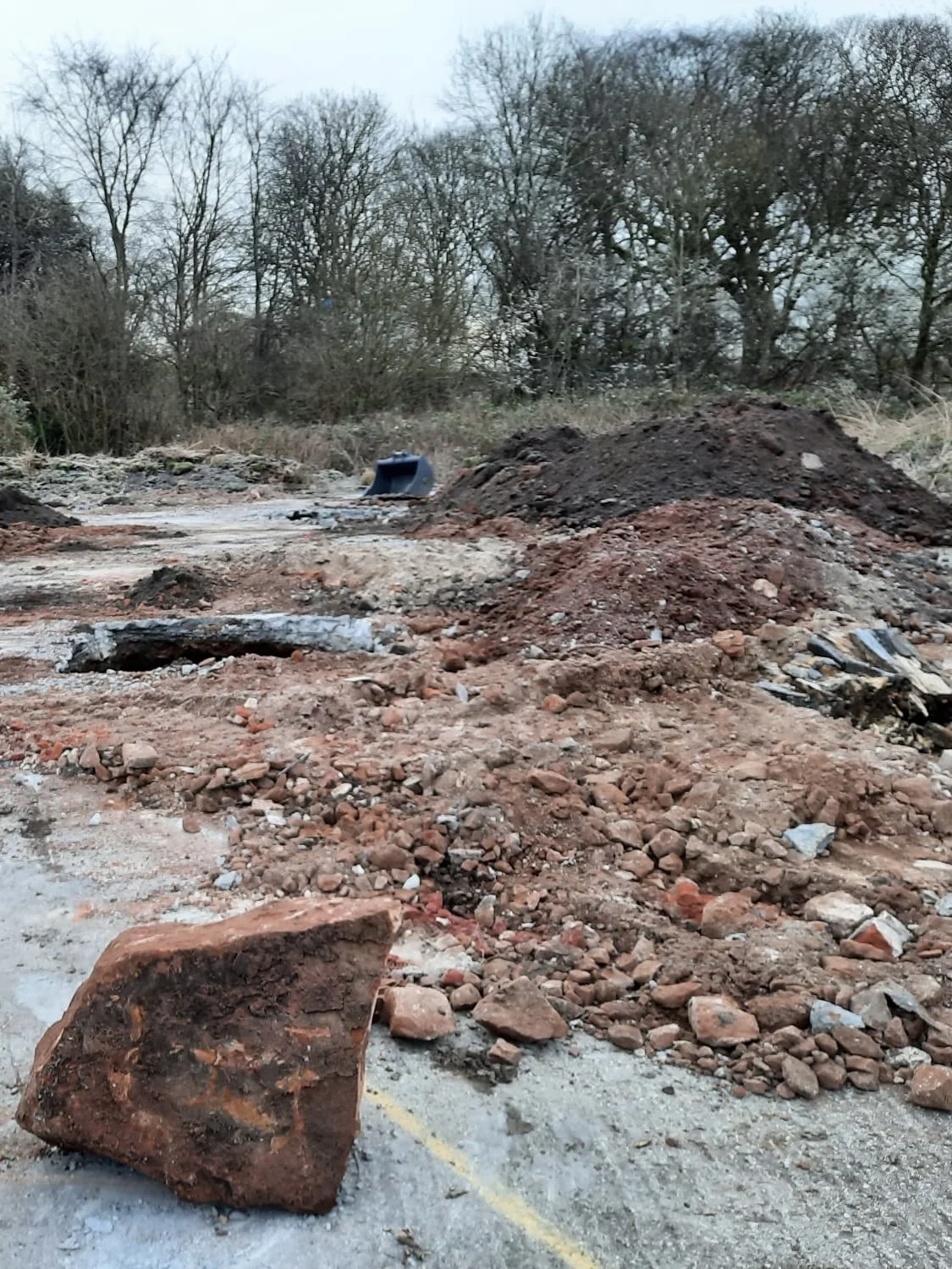Construction site with disturbed ground, soil and rocks, and a large boulder in the foreground; trees in the background.