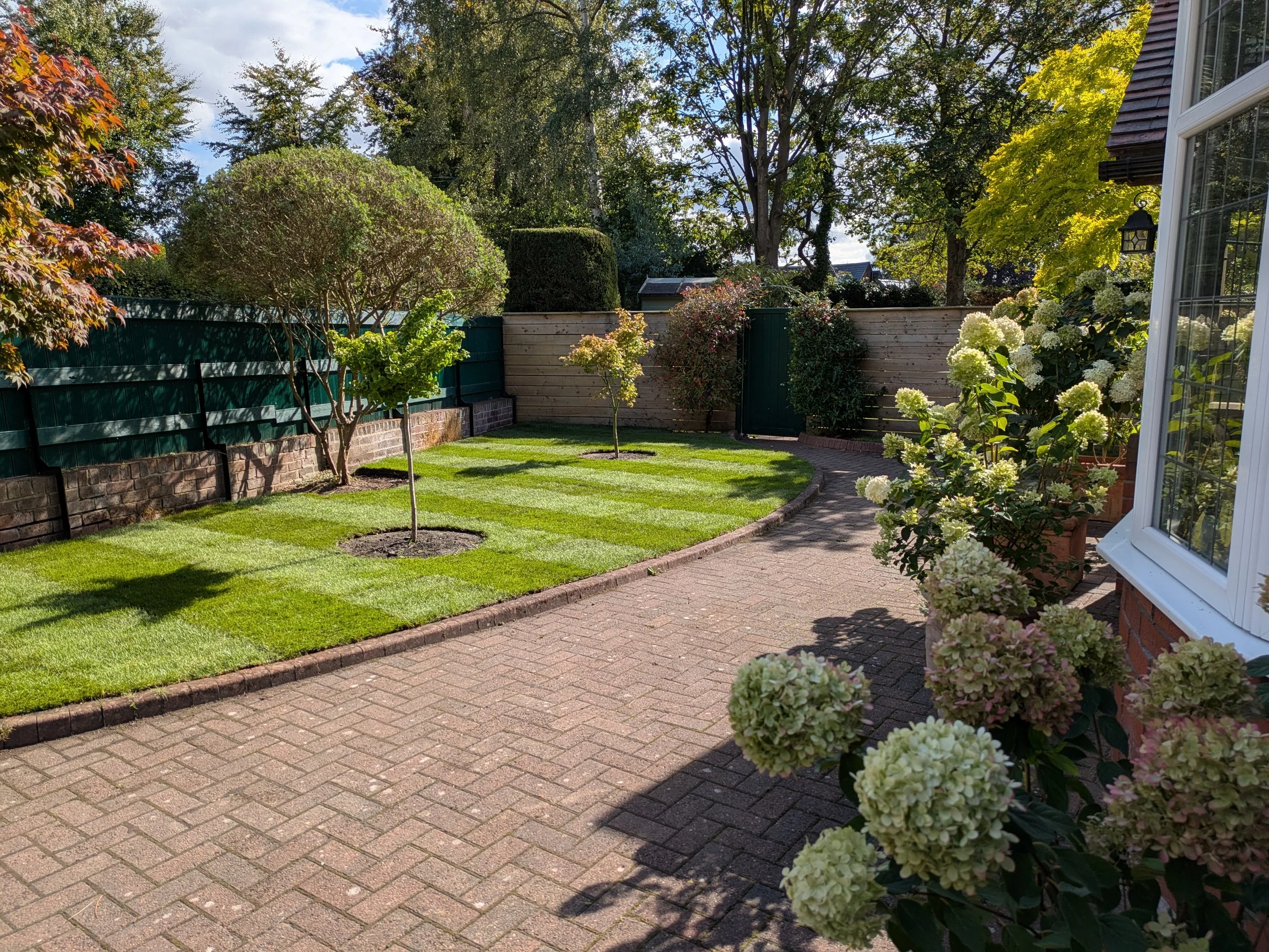 A well-maintained front garden with a brick patio, green grass, and small trees. There is a wooden fence, blossoming hydrangeas, and sunlight filtering through the trees.