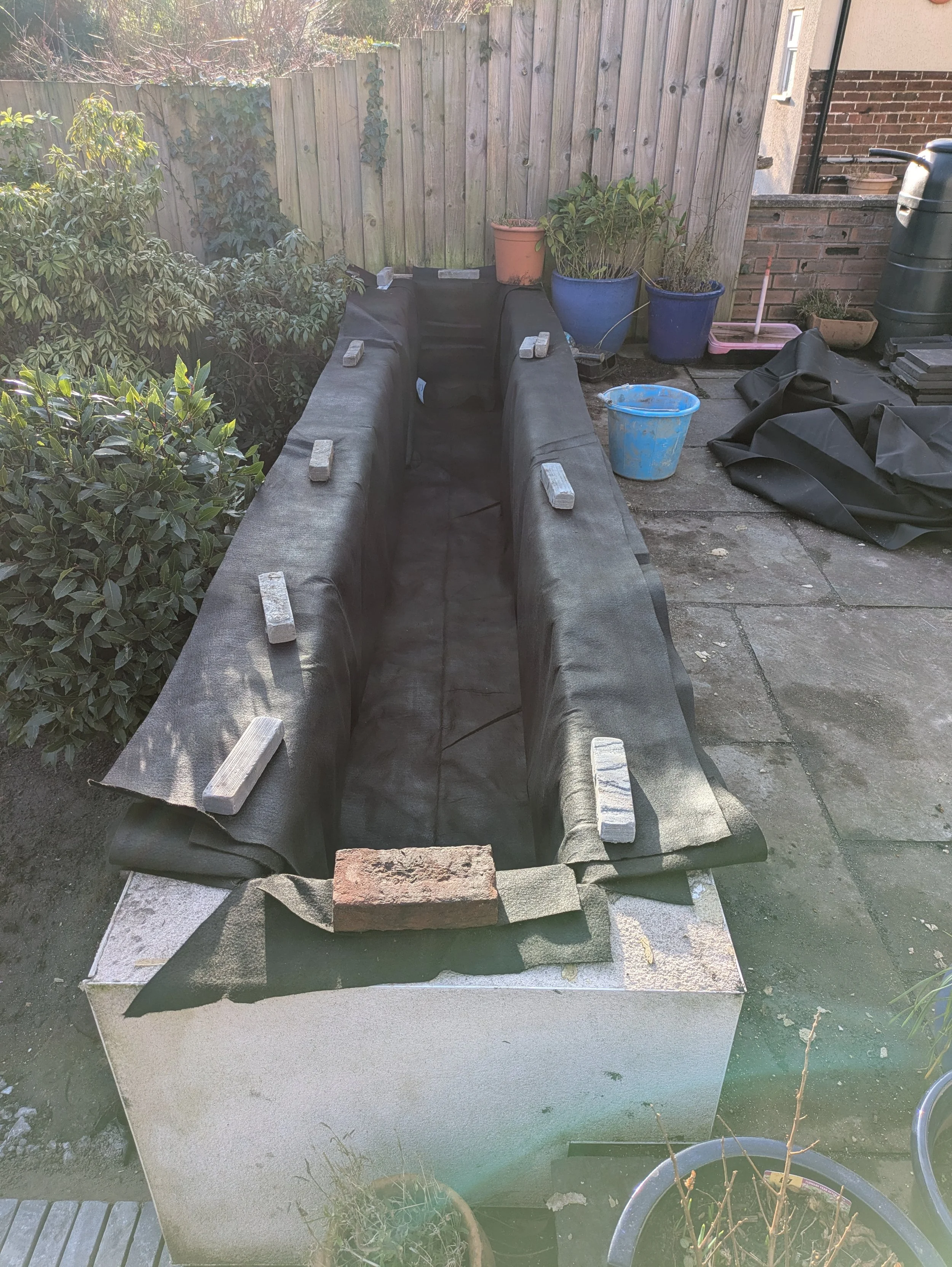 A partially assembled garden pond with a stone border, black liner, and small bricks inside, situated on a concrete patio. In the background are potted plants, a blue bucket, gardening supplies, and a wooden fence.