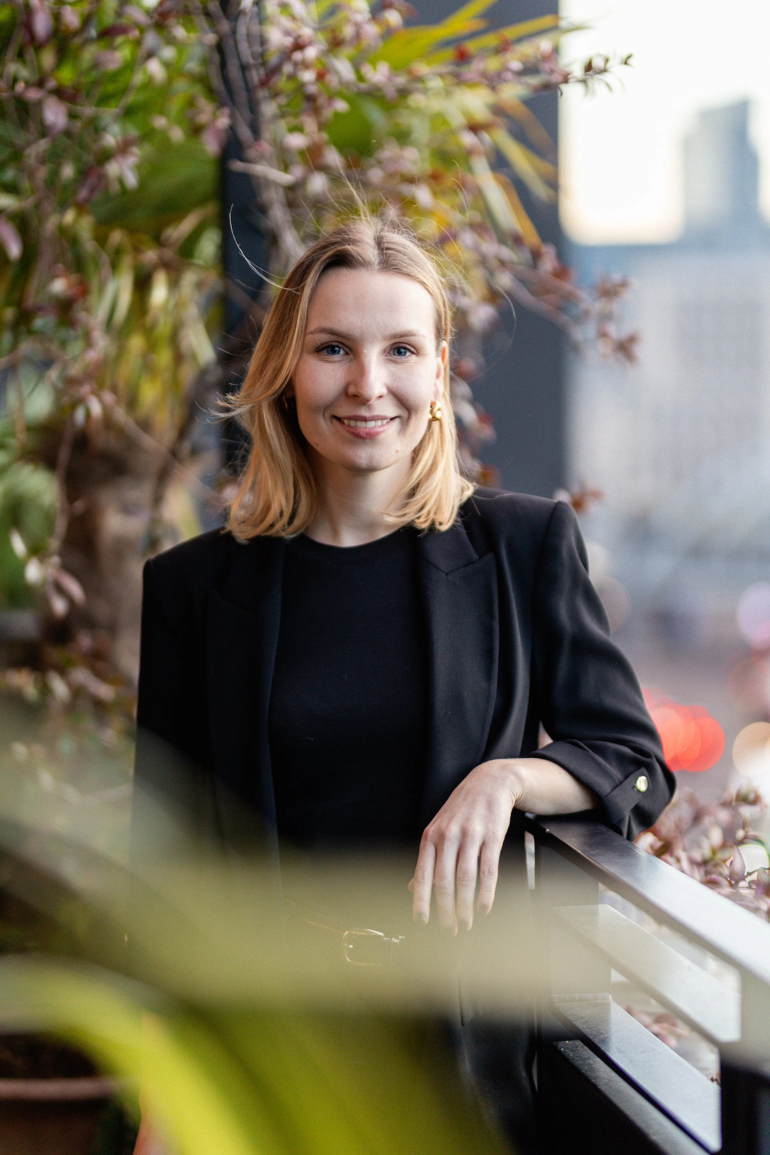 Beatrice Grabazyte: A young woman with blond hair smiling, wearing a black blazer and top, standing on a balcony with pink flowers and city buildings in the background.