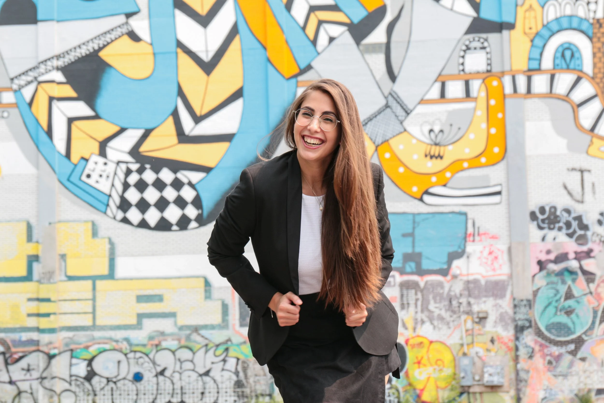 Fadia Frangie Doumat: Smiling woman with long hair and glasses wearing a black blazer and skirt, standing in front of a colorful graffiti wall.
