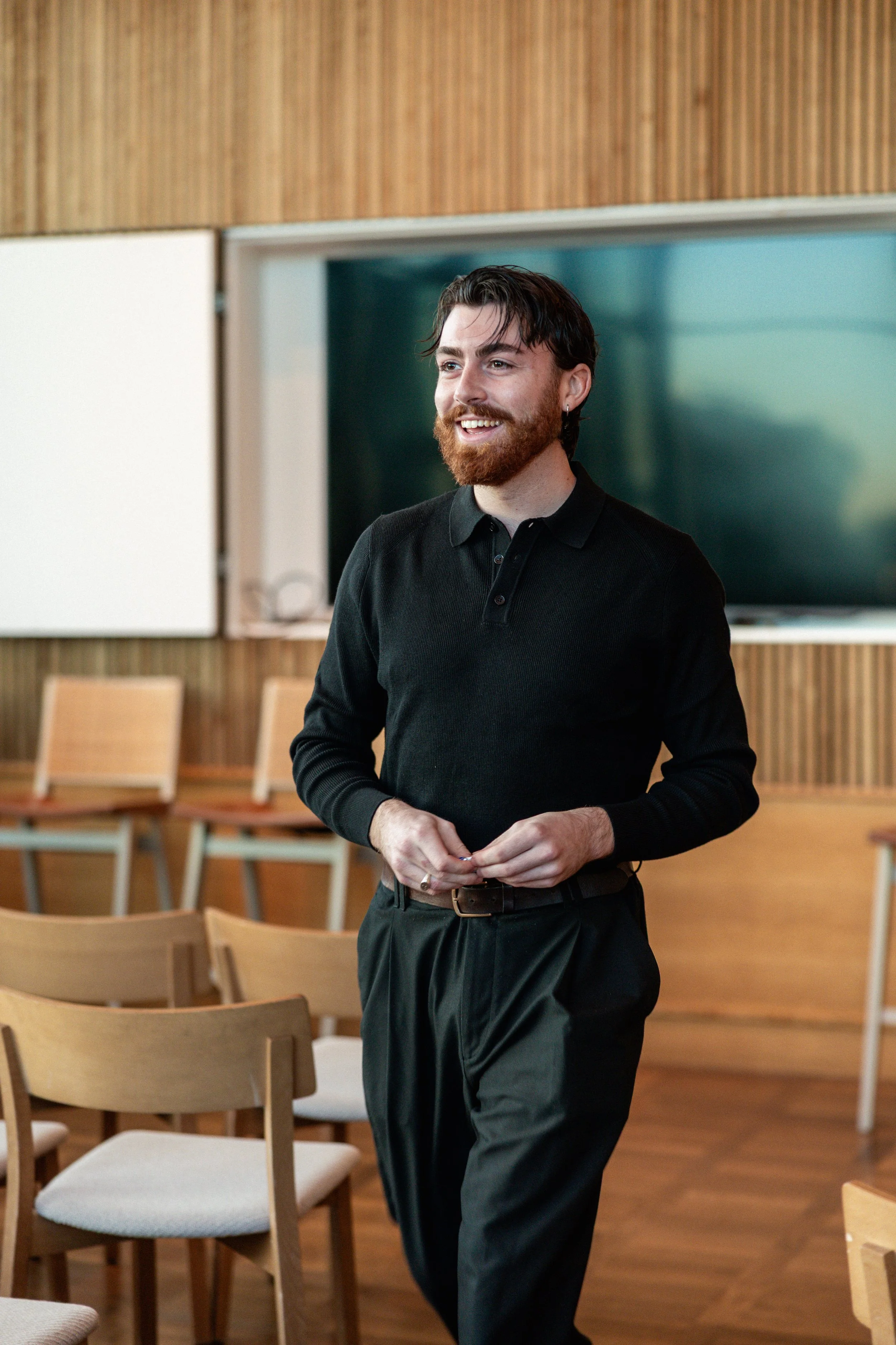 Sean Harris: A man with dark hair and a beard smiling, standing in a conference room with wooden chairs, a large TV screen, and wood paneling on the wall.