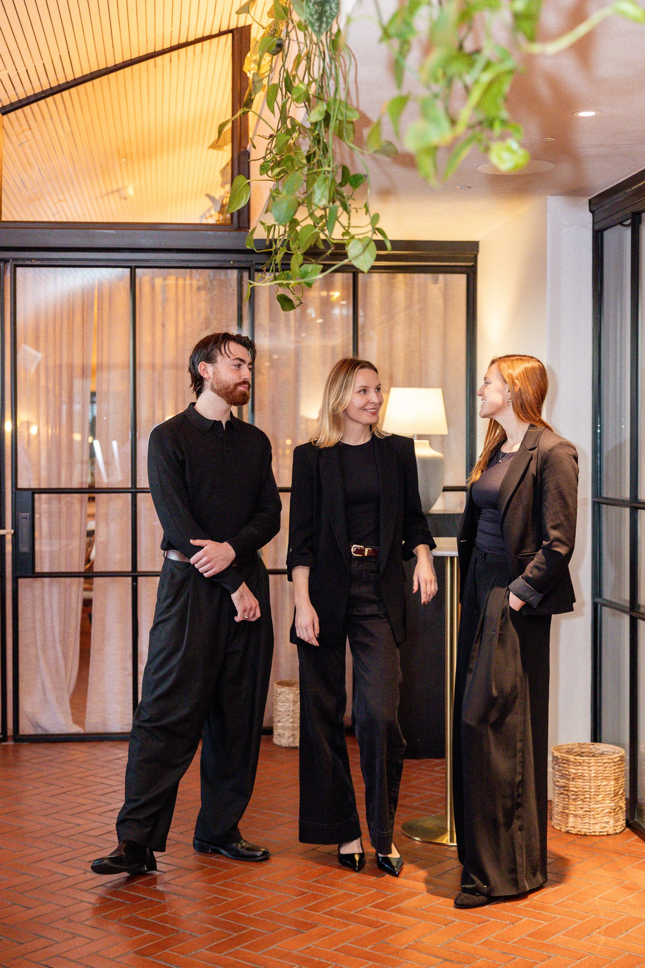 Three people in black formal attire engaging in conversation inside a restaurant with a brick floor and warm lighting.
