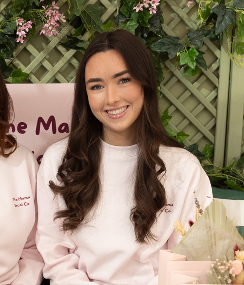 Evie Doman: Young woman with long dark hair and a white sweatshirt smiling at a social event with pink and floral decorations.